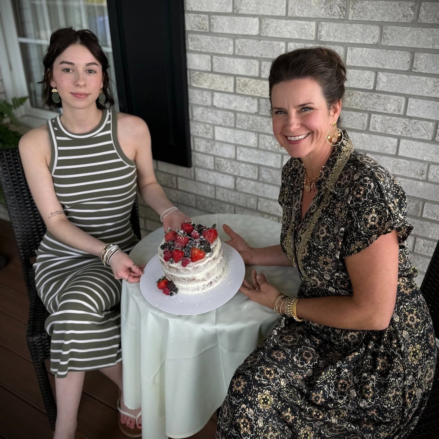 Two people seated by a table with a white cake topped with red flowers, smiling indoors.