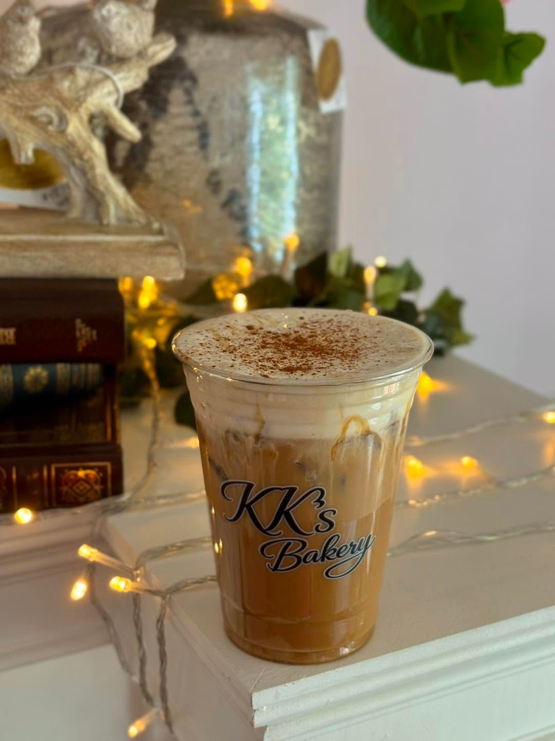 Iced coffee in a clear plastic cup with “KK’s Bakery” logo on a marble table with warm fairy lights.