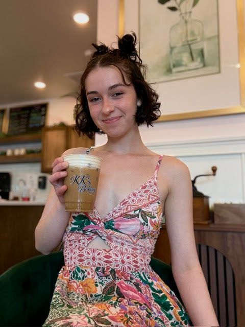 Woman in a floral dress holding an iced drink in a café, smiling at the camera.