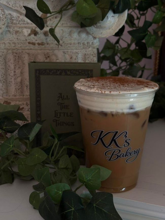 Iced coffee in a clear cup with whipped foam, on a tray with green leaves and a decorative sign.