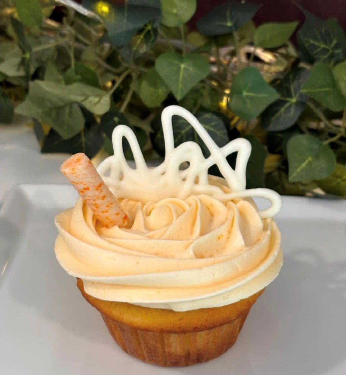 Vanilla cupcake with pale frosting, white drizzle, and a wafer curl on a white tray, with green leaves behind