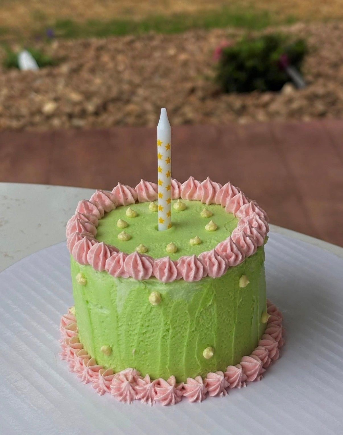 Green frosted birthday cake with pink piping and a single striped candle on a table outdoors