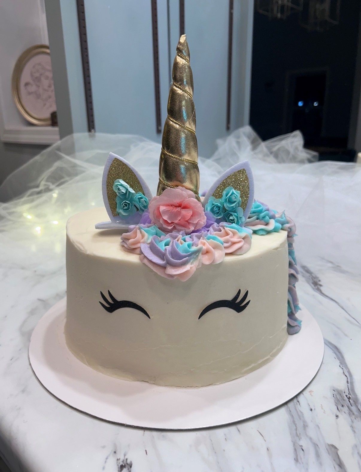 Unicorn-themed white cake with gold horn, pastel flowers, and closed-eye face design on a white stand