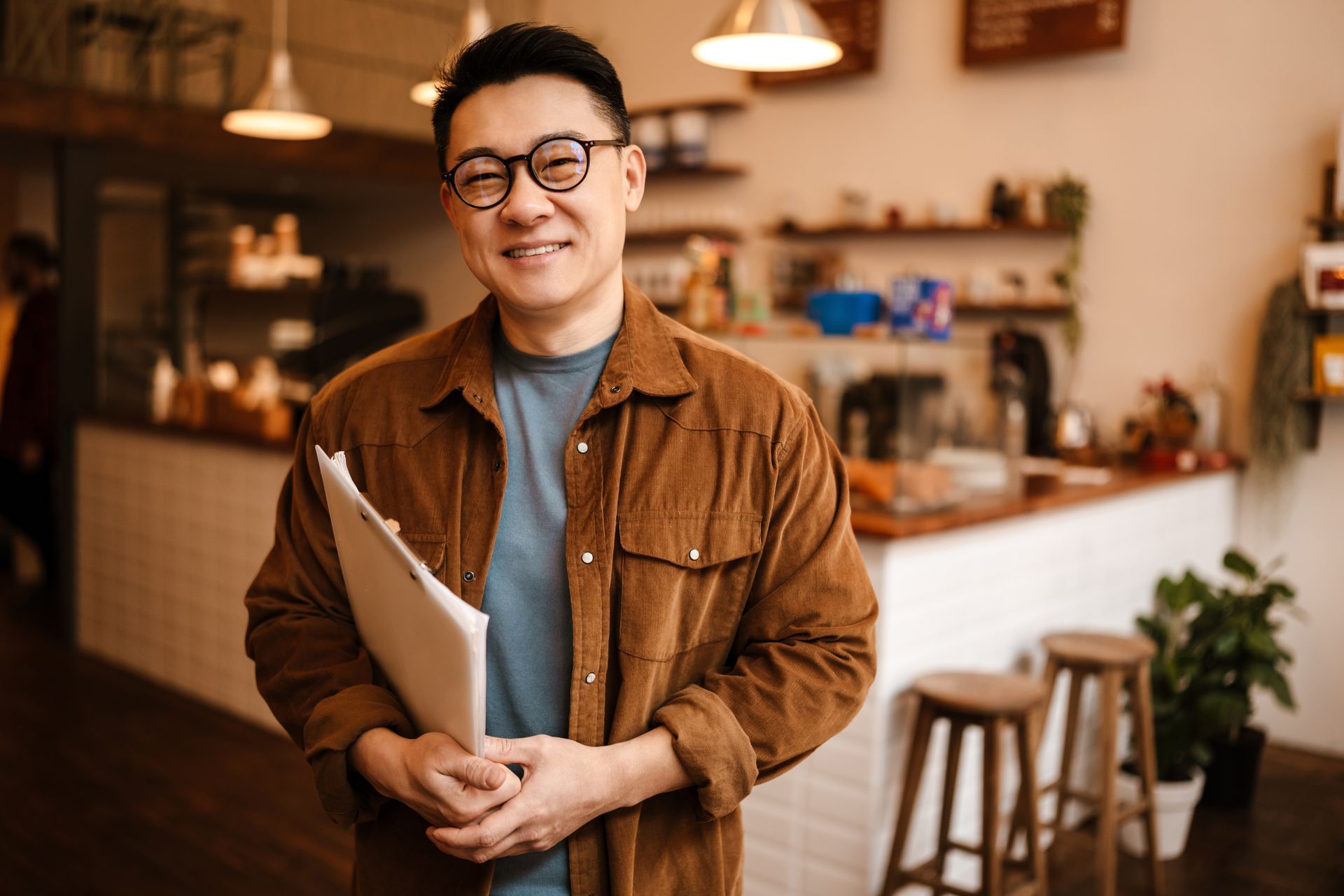 Un homme portant des lunettes et une chemise en velours côtelé tient des papiers et sourit dans un café avec des tabourets et un comptoir.