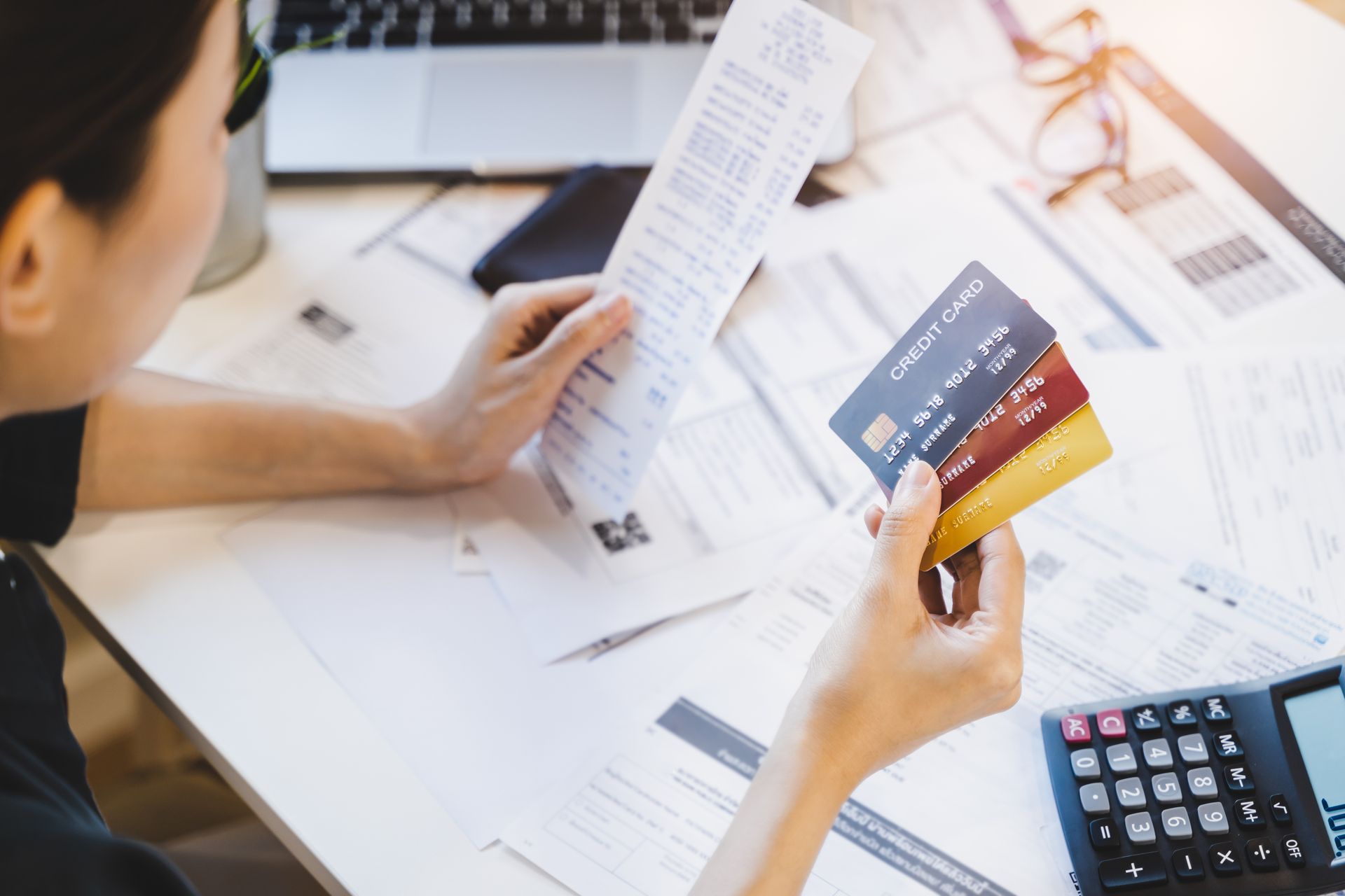 Une femme examine des factures, tenant des cartes de crédit, assise à un bureau encombré avec une calculatrice et un ordinateur portable.