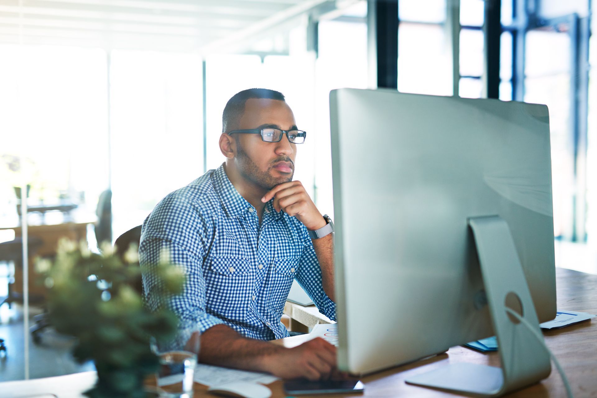 Un homme portant des lunettes regarde attentivement un écran d'ordinateur dans un bureau lumineux.