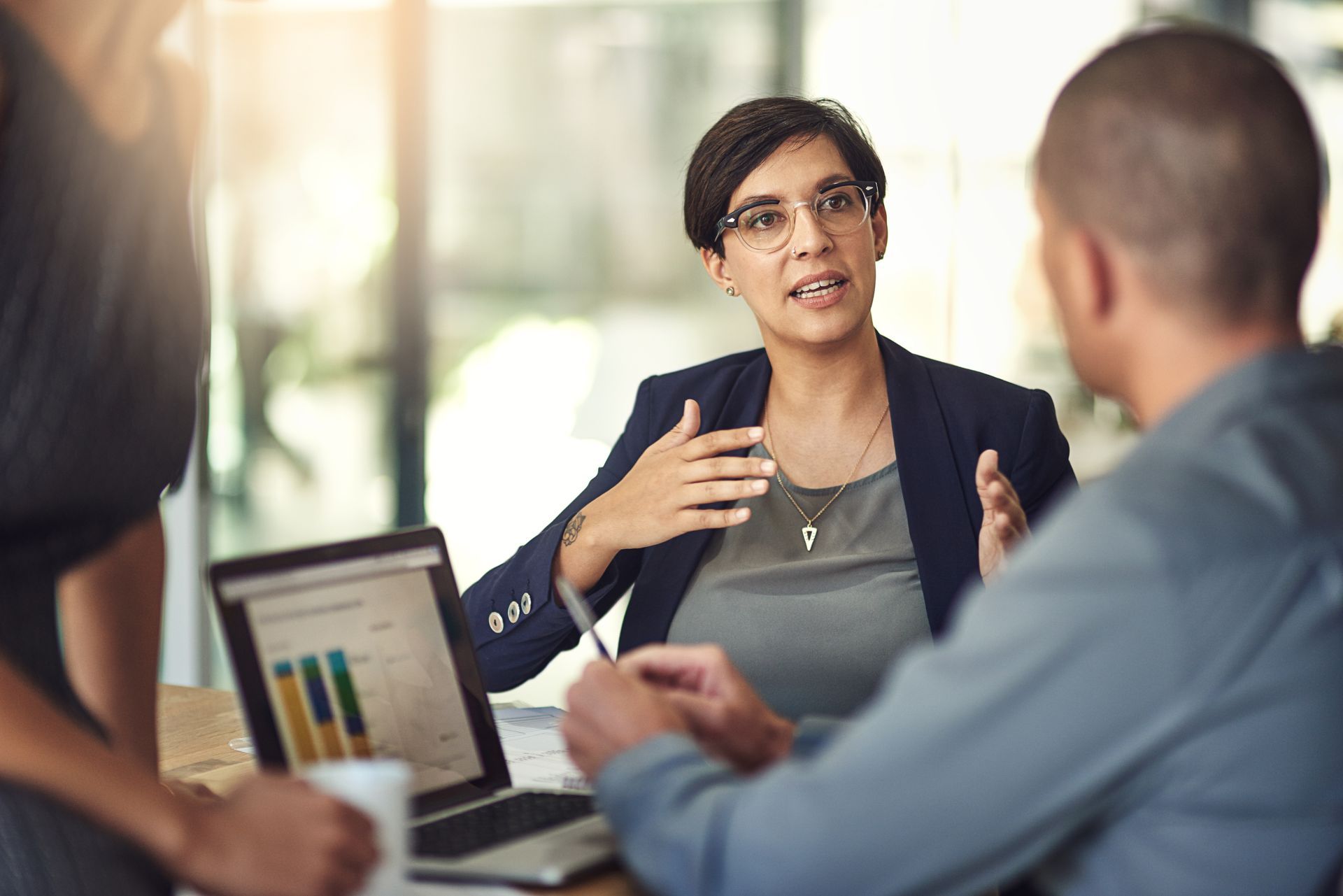 Femme en blazer faisant des gestes tout en discutant de données avec des collègues autour d'une table ; ordinateur portable ouvert.