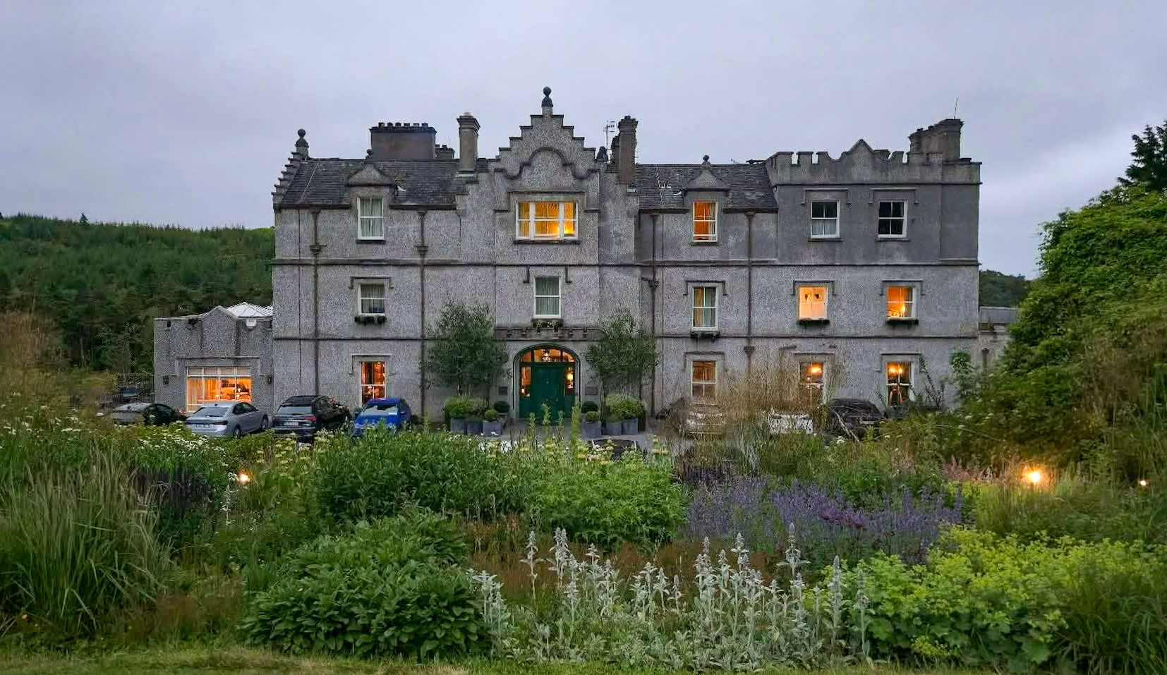 Stone manor house at dusk, lit windows glowing above a lush garden and overgrown greenery.