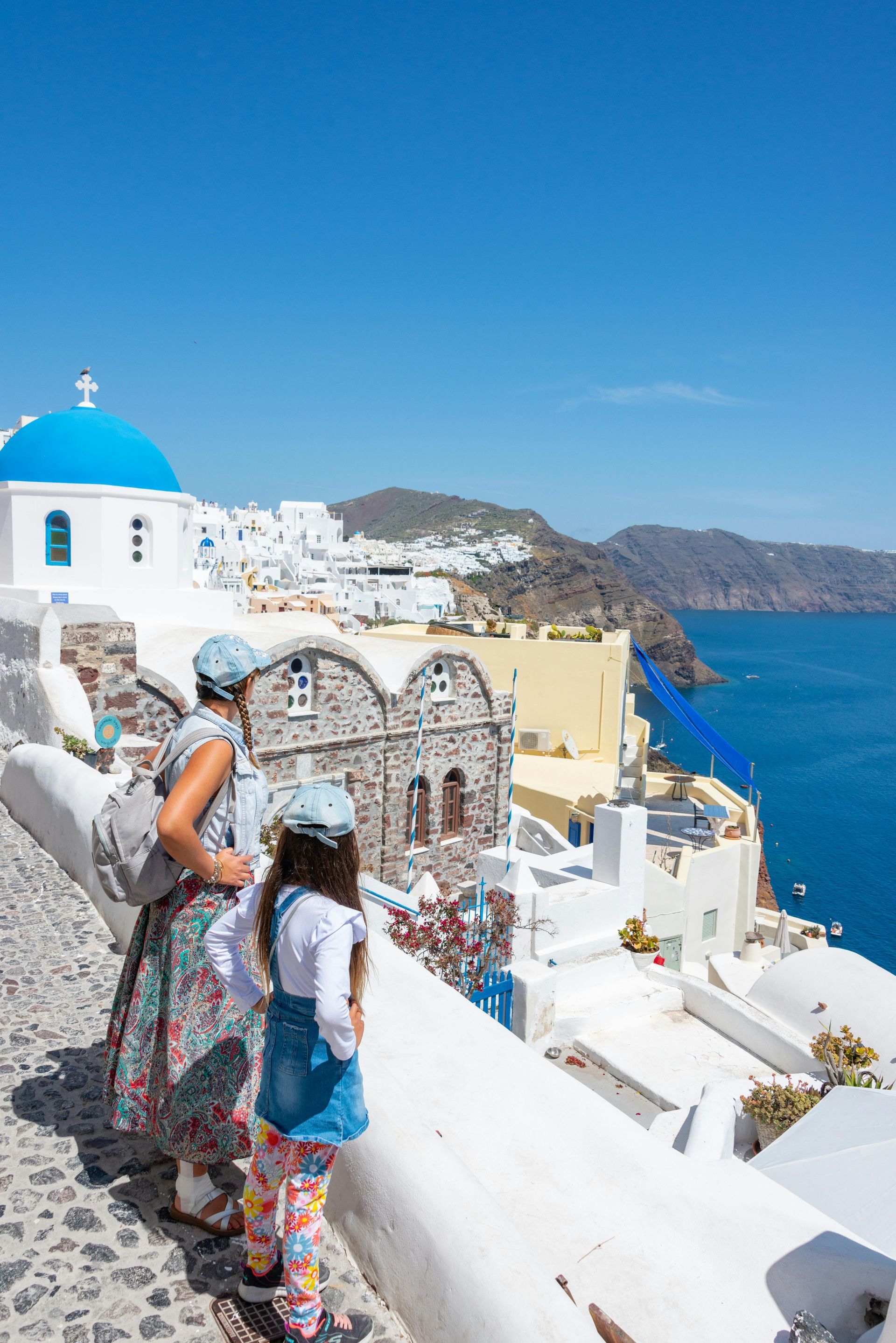 Tourists overlooking a white hillside village with blue-domed church and bright Aegean sea.