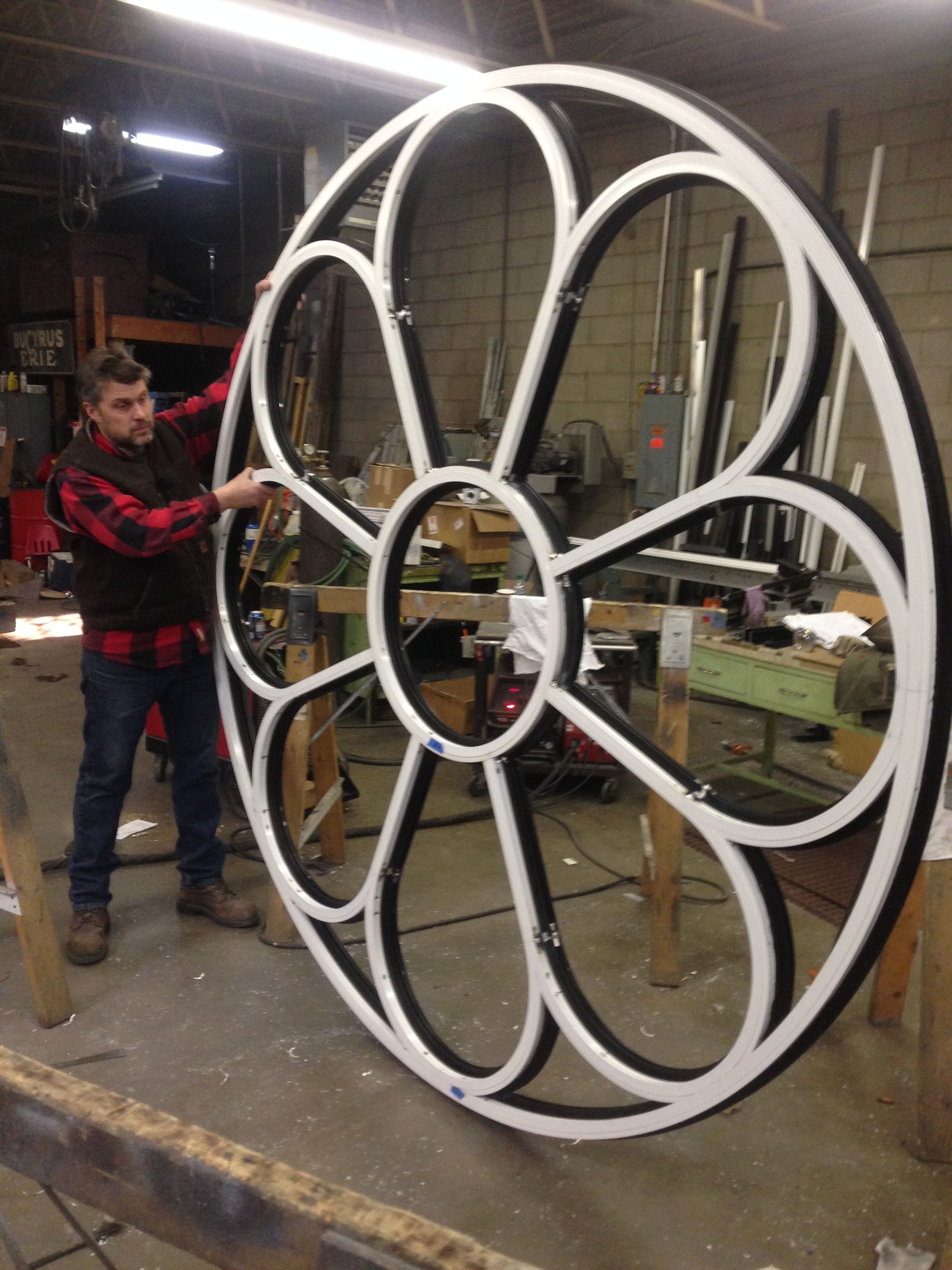 A man in a plaid vest is holding a large black and white wheel in a workshop.