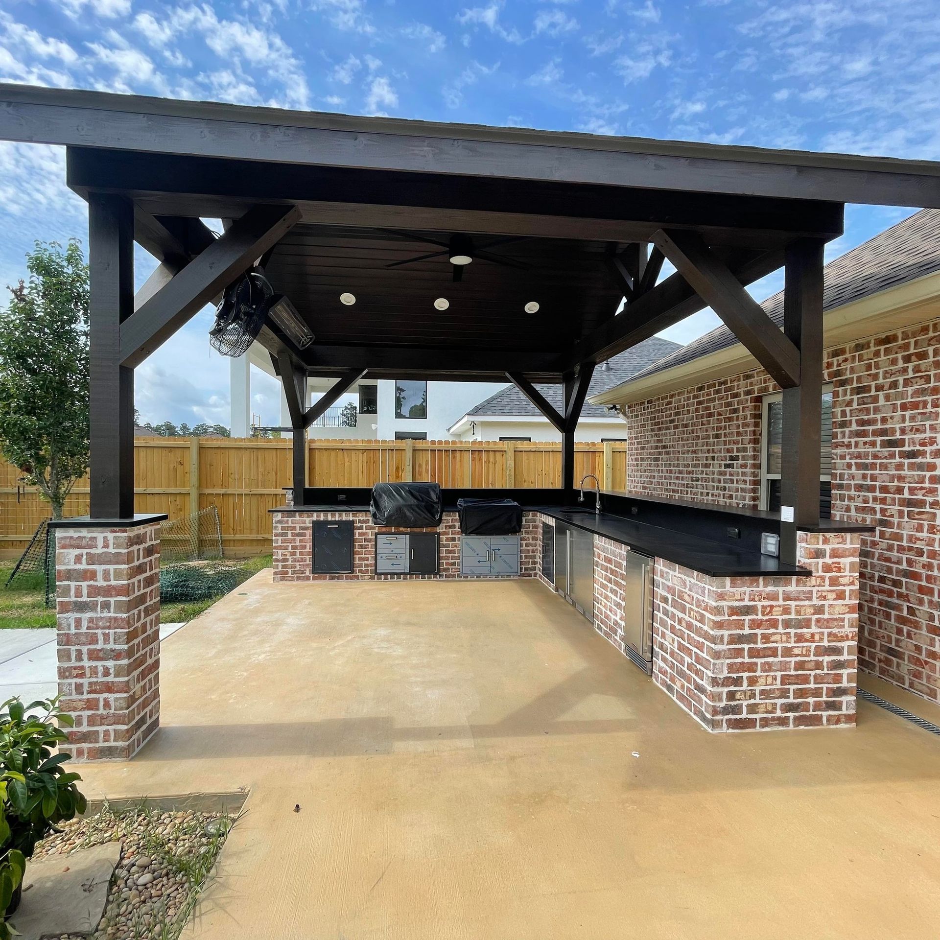 An outdoor kitchen with a brick base and dark stone countertop under a covered wooden patio structure.