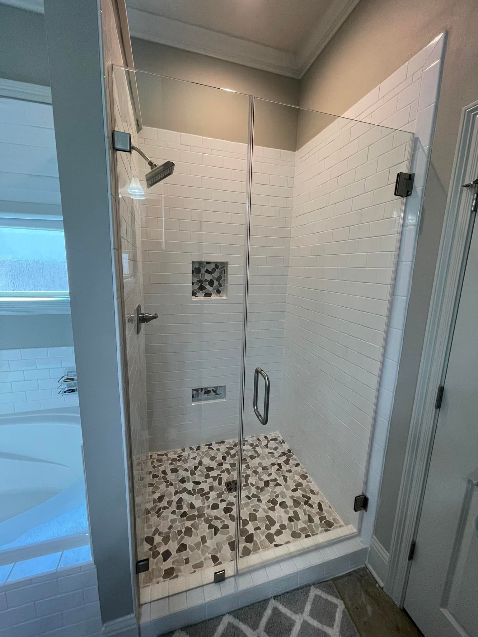 A walk-in shower with white horizontal tiled walls, a pebble-stone floor, a glass door, and a built-in shelf niche.