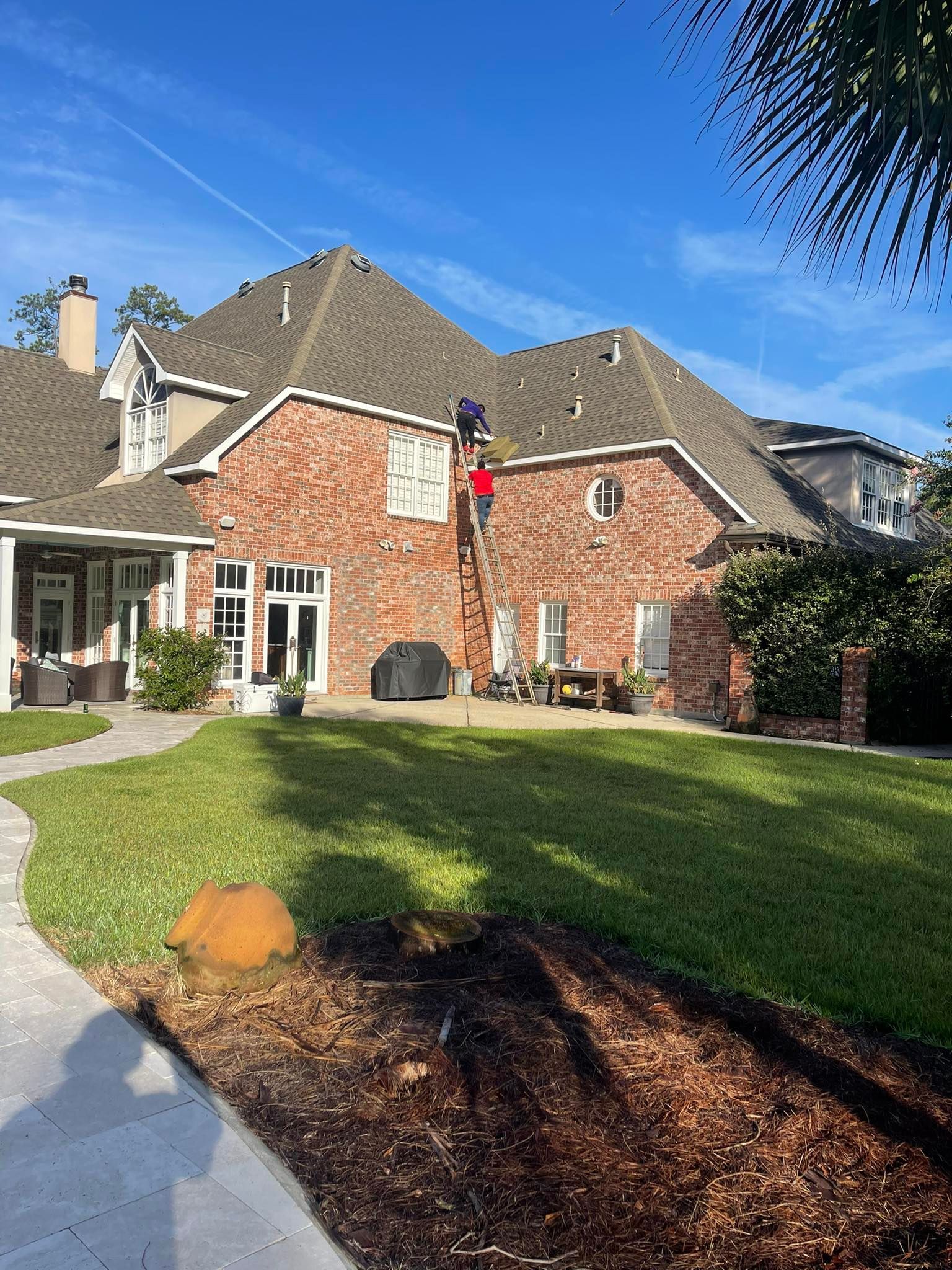 Two people in red shirts use a ladder to work on the roof of a large brick house with a grassy backyard.