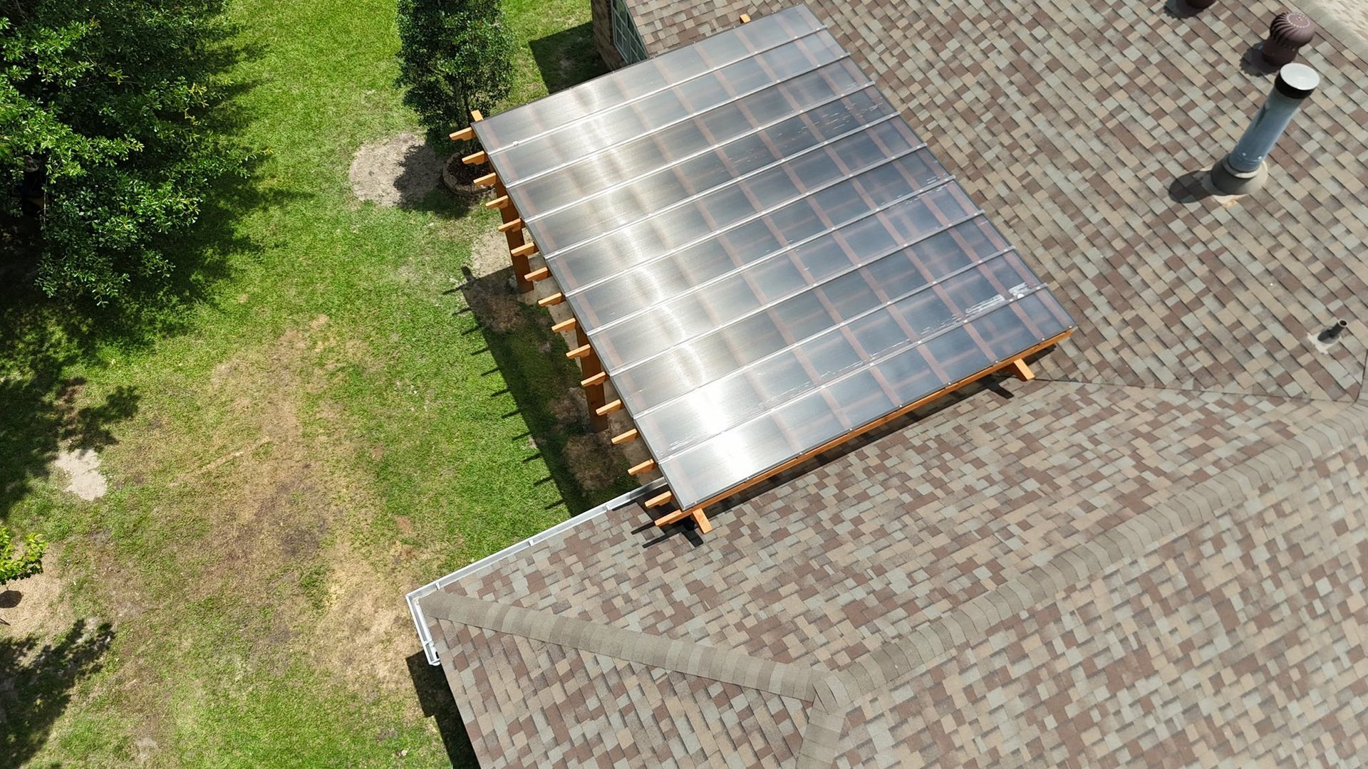 An aerial view shows a solar panel array mounted on a wooden frame attached to the shingled roof of a house near a lawn.