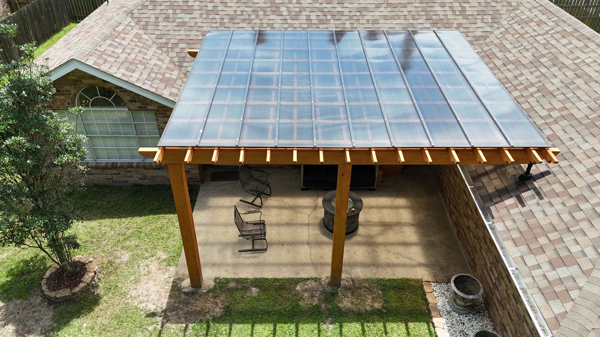 An elevated view of a wooden patio pergola with solar panels installed on its roof, adjacent to a brick house.