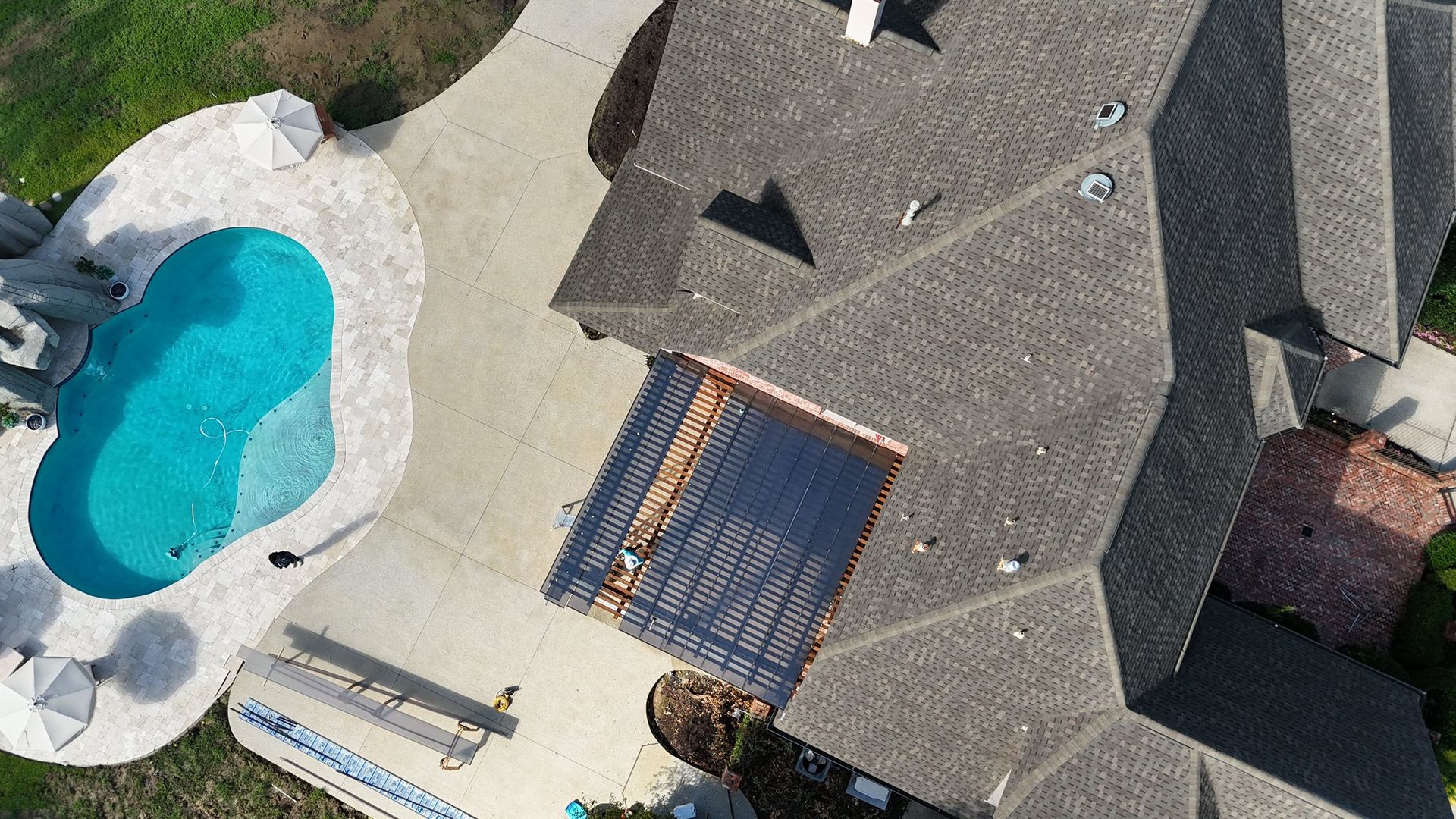 An aerial view of a house with a gray shingled roof, a patio with a wooden pergola, and a turquoise kidney-shaped pool.