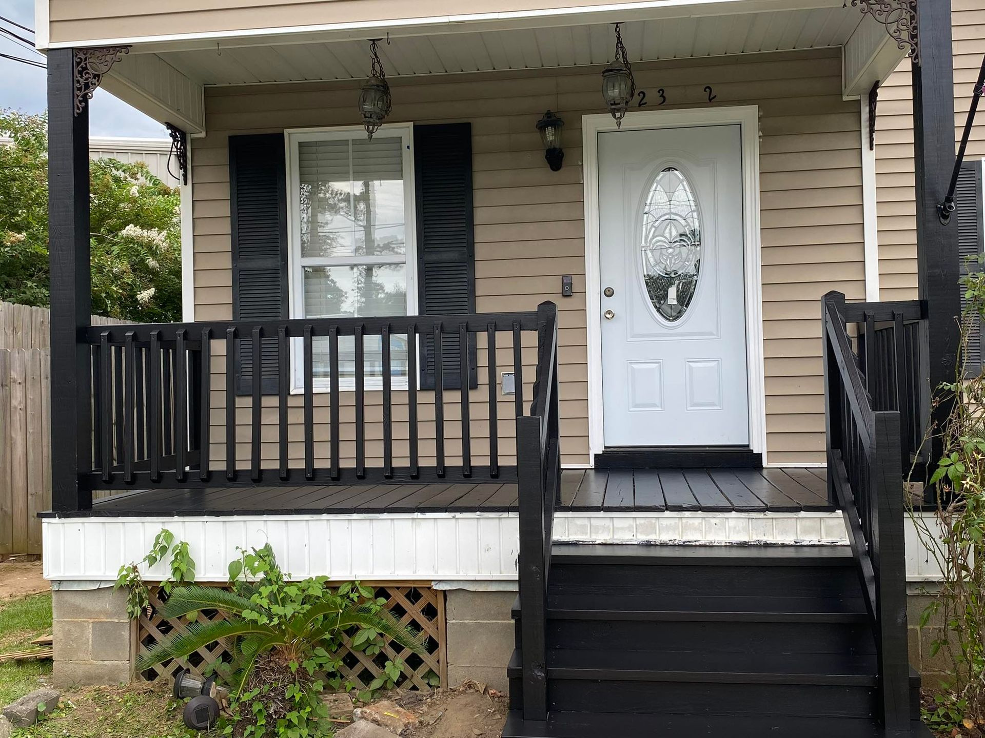 A tan house porch with black railings and steps, a white door with an oval glass window, and a white lattice base.