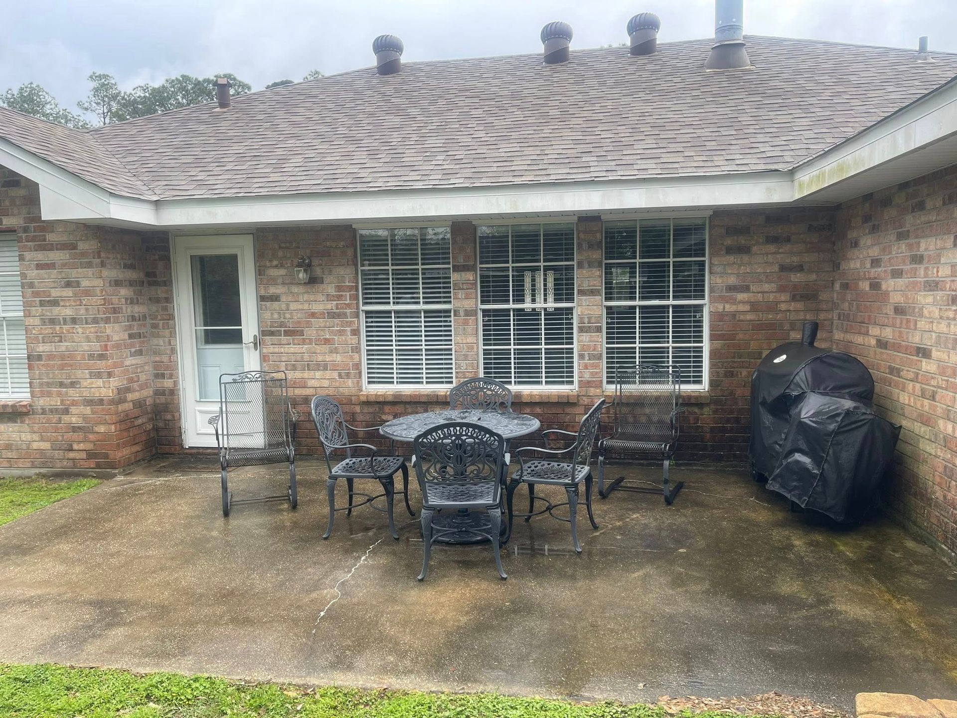 A patio with a round metal table, four matching chairs, and a covered grill against a brick house wall.