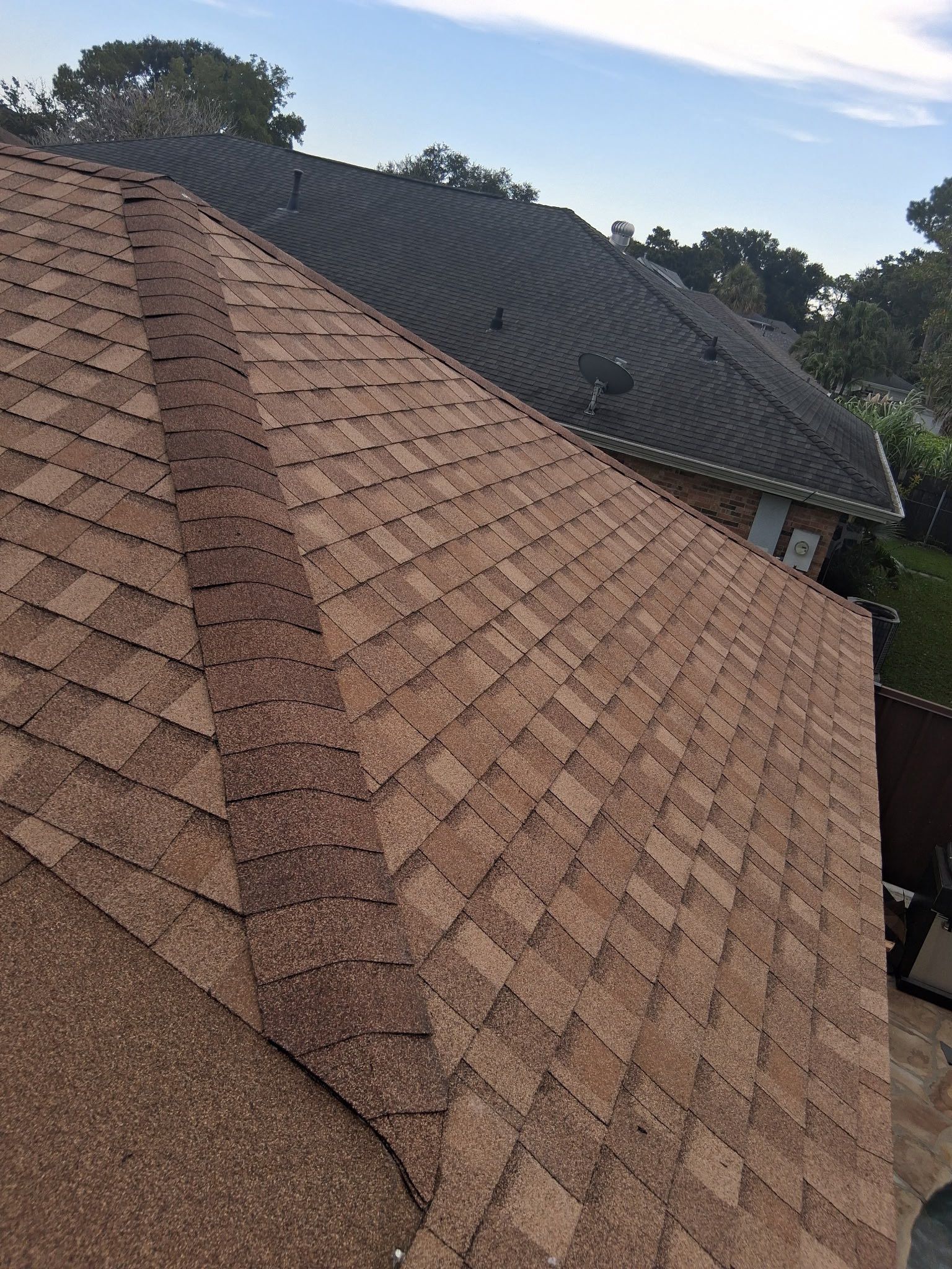 An angled view of a roof with tan asphalt shingles and a distinct ridge cap, with a darker roof visible in the background.