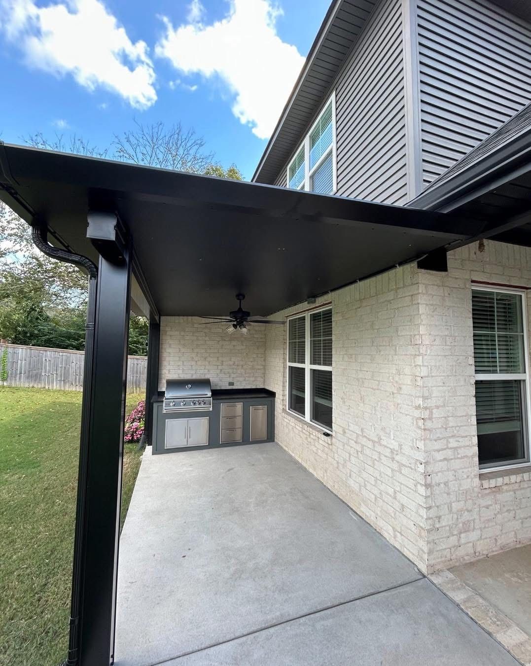 A covered brick patio area featuring an outdoor grill station beneath a black awning with a view of a grassy lawn.