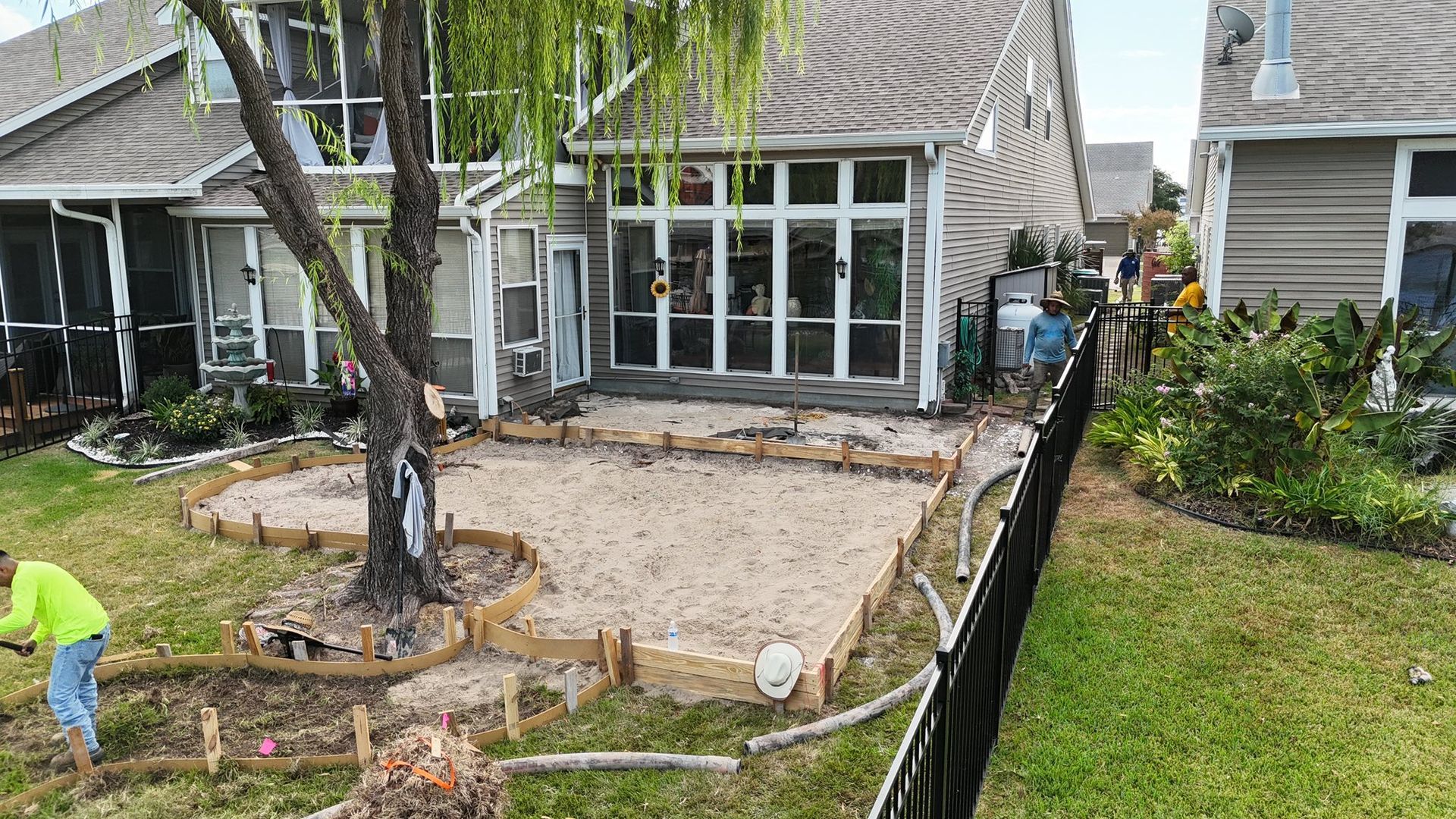 A worker installs wooden forms for a patio foundation in a residential backyard with a large tree.