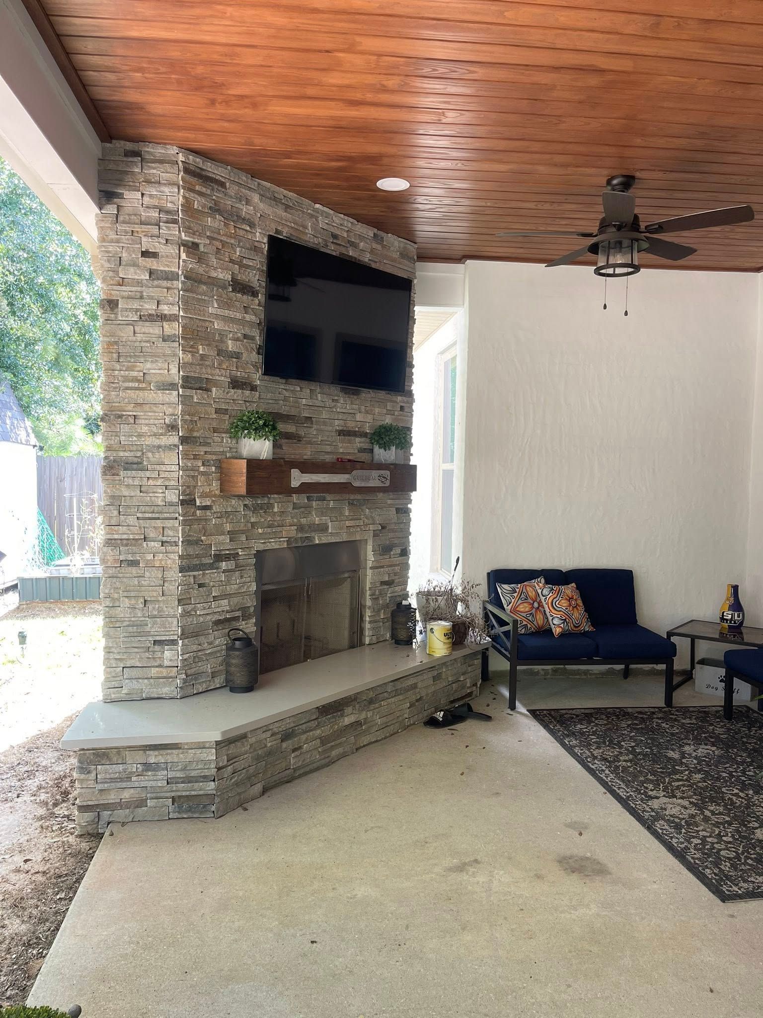 A stone fireplace with a mounted television and wood mantel on a covered outdoor patio with a blue sofa and rug.