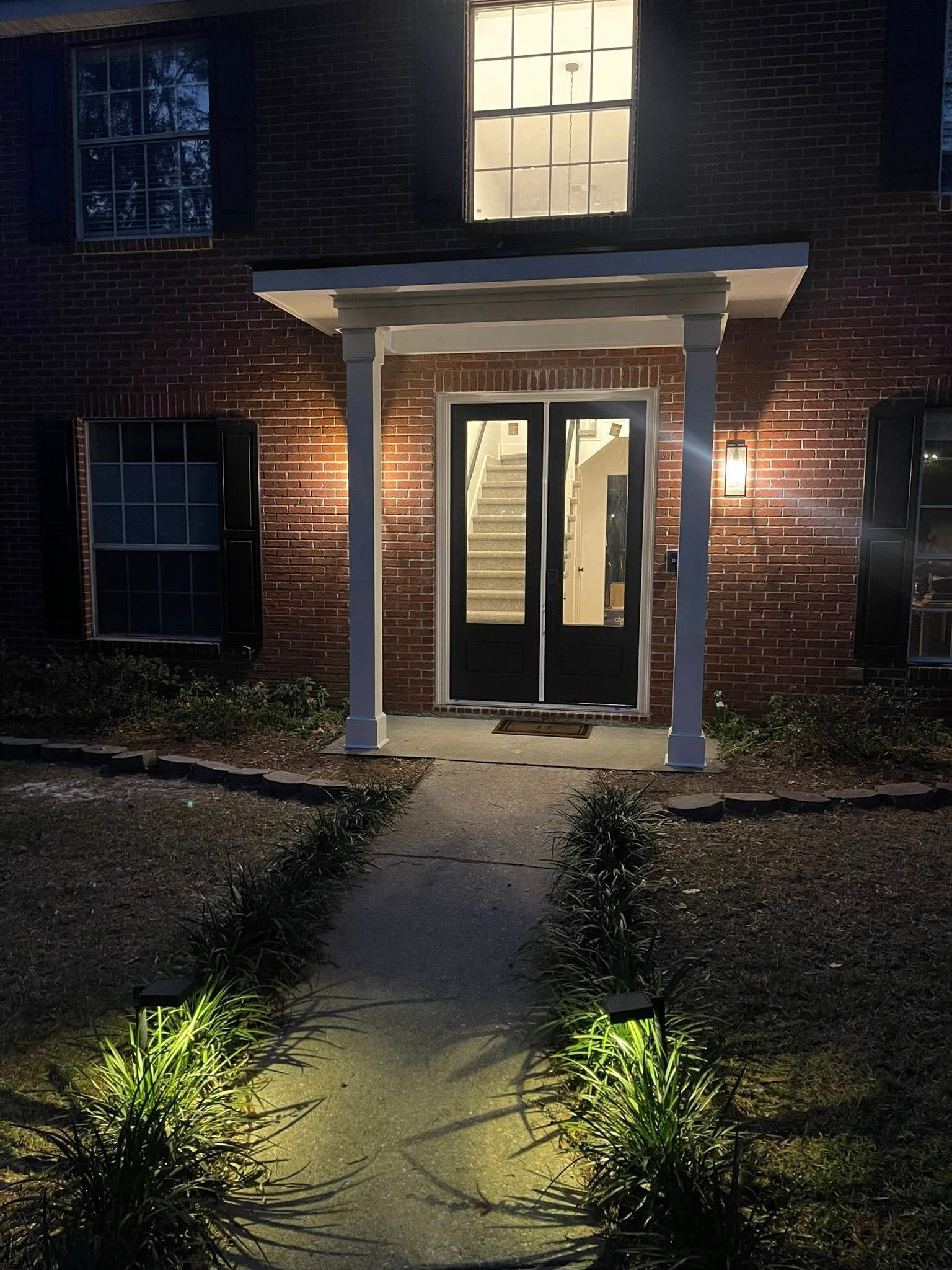 A night view of a brick house entrance with a lit path, white porch columns, and a view into the bright foyer.