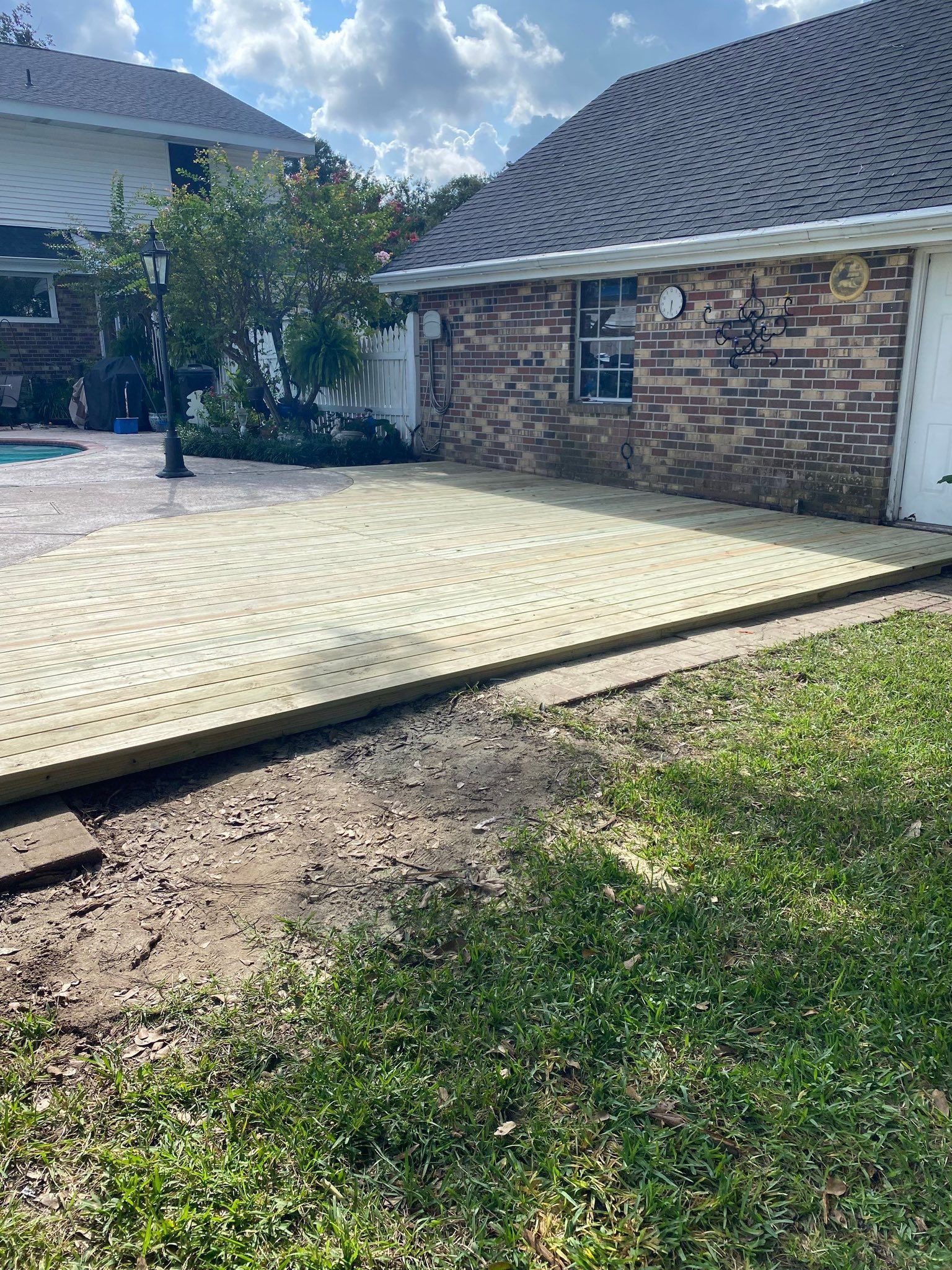 Newly installed light-colored wooden deck in a backyard, adjacent to a brick house exterior and a pool area.