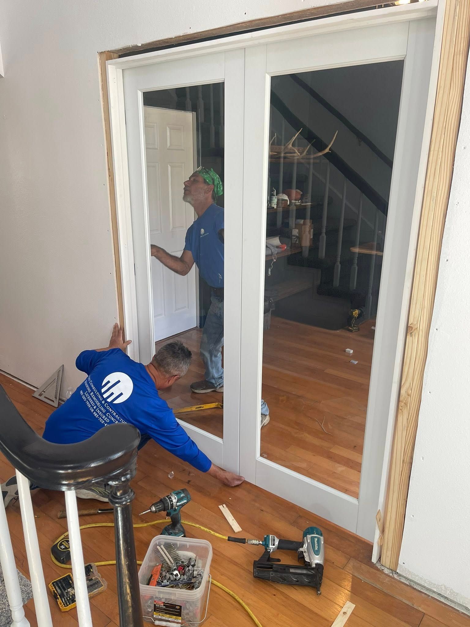 Two workers in blue shirts install a double door in a residential home, with power tools visible on the wooden floor.