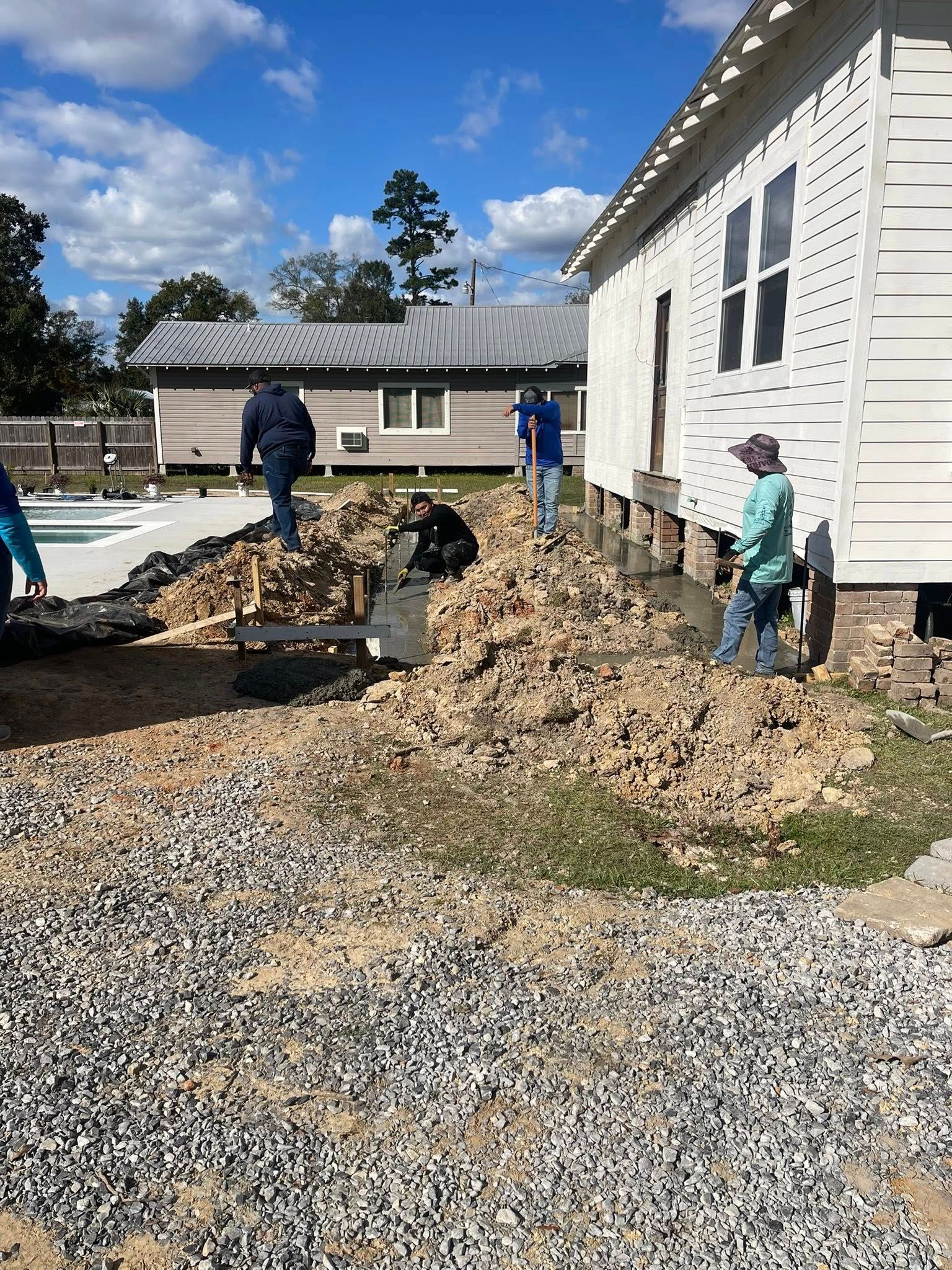 Workers perform foundation construction at a white building site, digging a trench in dirt alongside gravel ground.