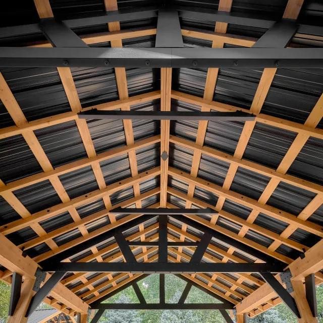 An open-air pavilion ceiling showing a structure of natural wood rafters, black metal beams, and a dark corrugated roof.
