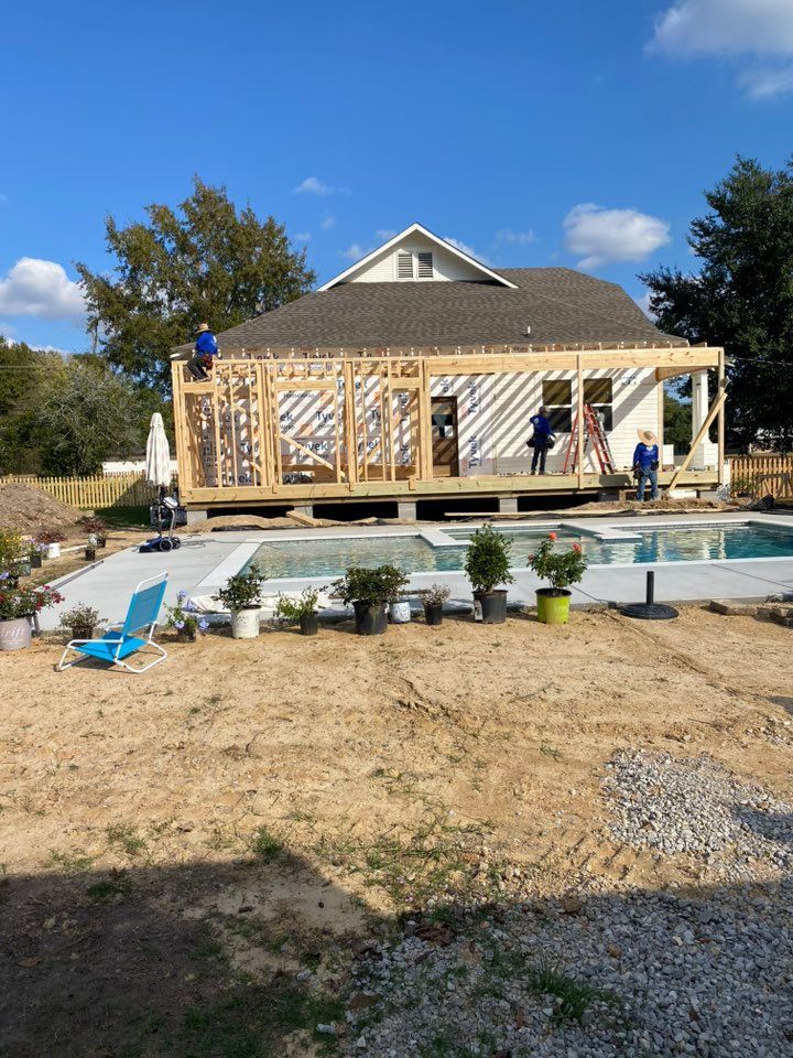 Construction crew working on a wooden patio frame attached to a house behind a swimming pool in a sunny backyard.