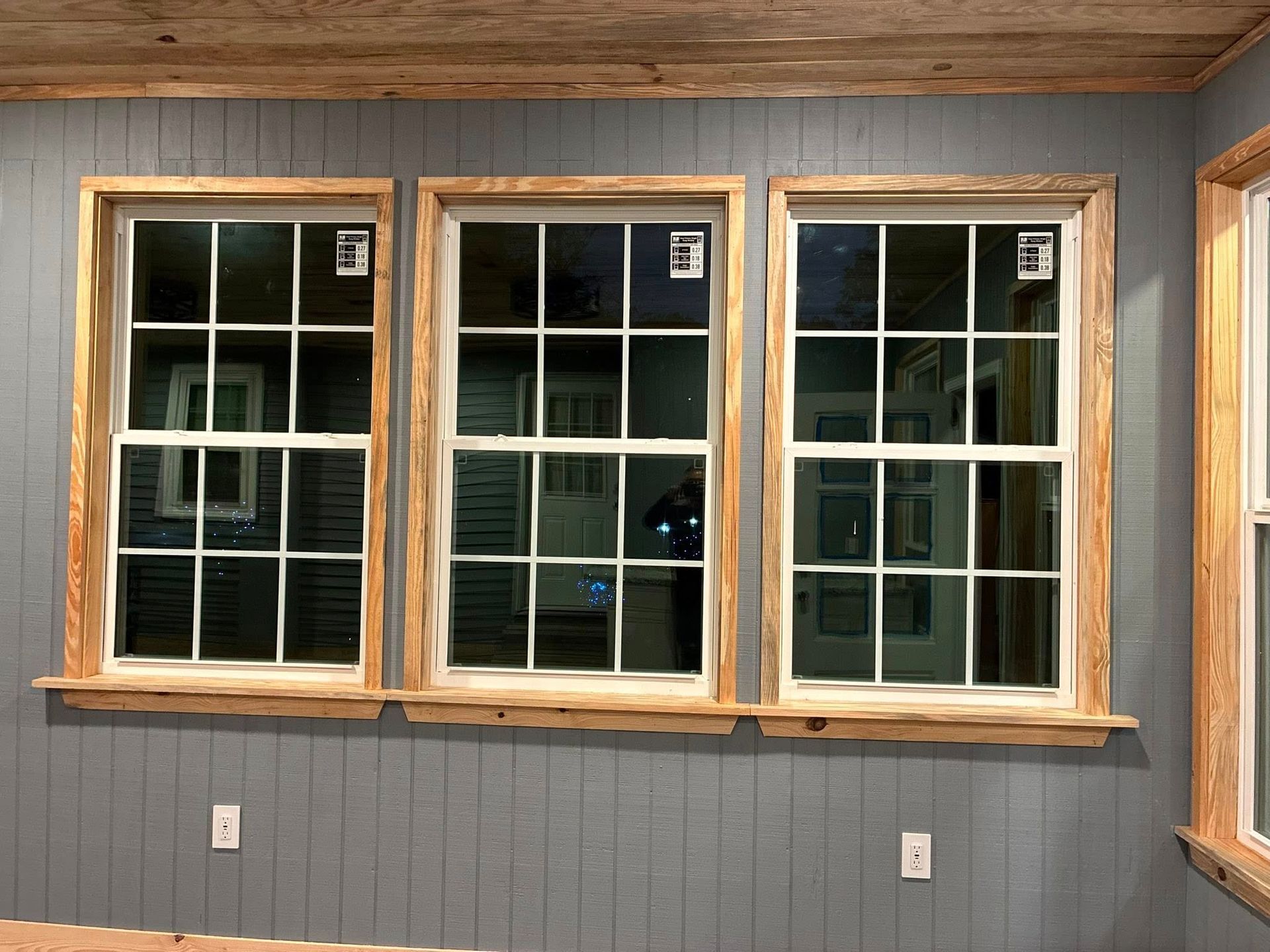 Three white-paned windows with rustic wooden frames set into a grey vertical-planked wall with two white wall outlets.