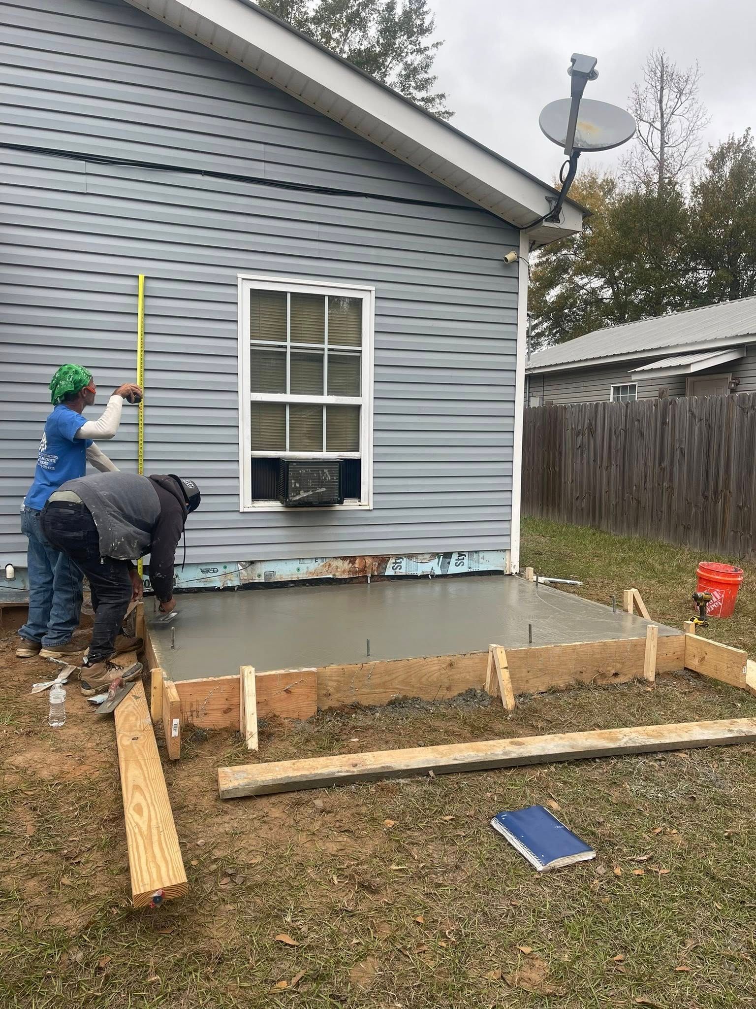 Construction workers pour a concrete pad in front of a blue-sided house while measuring the wall height.
