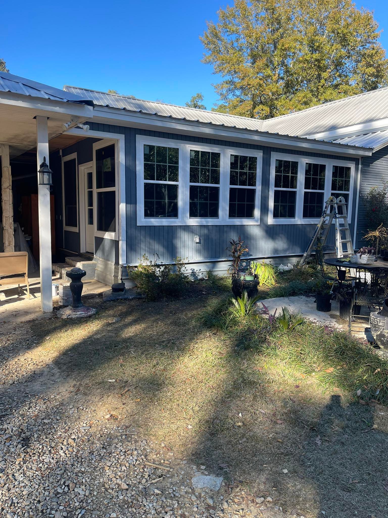 A blue-sided house extension with a row of white-framed windows, a covered porch, and a gravel yard under a blue sky.