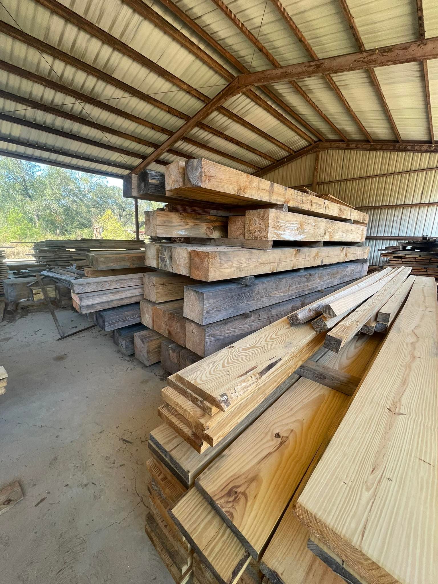 Stacks of rough-cut wooden planks and large beams stored inside an open-air metal shed.