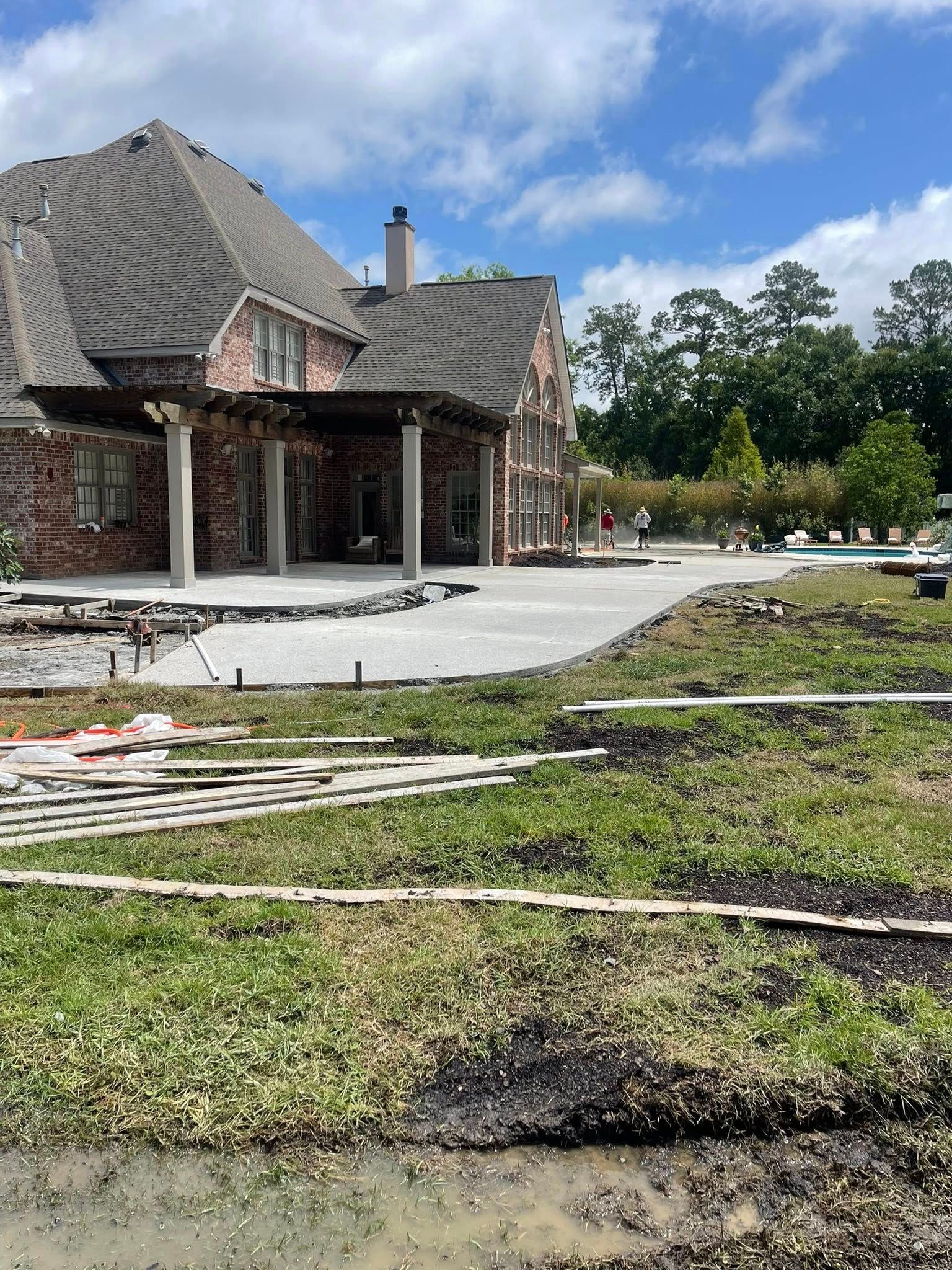 A brick house with a newly poured concrete patio and pergola in a backyard with patchy grass and muddy soil.