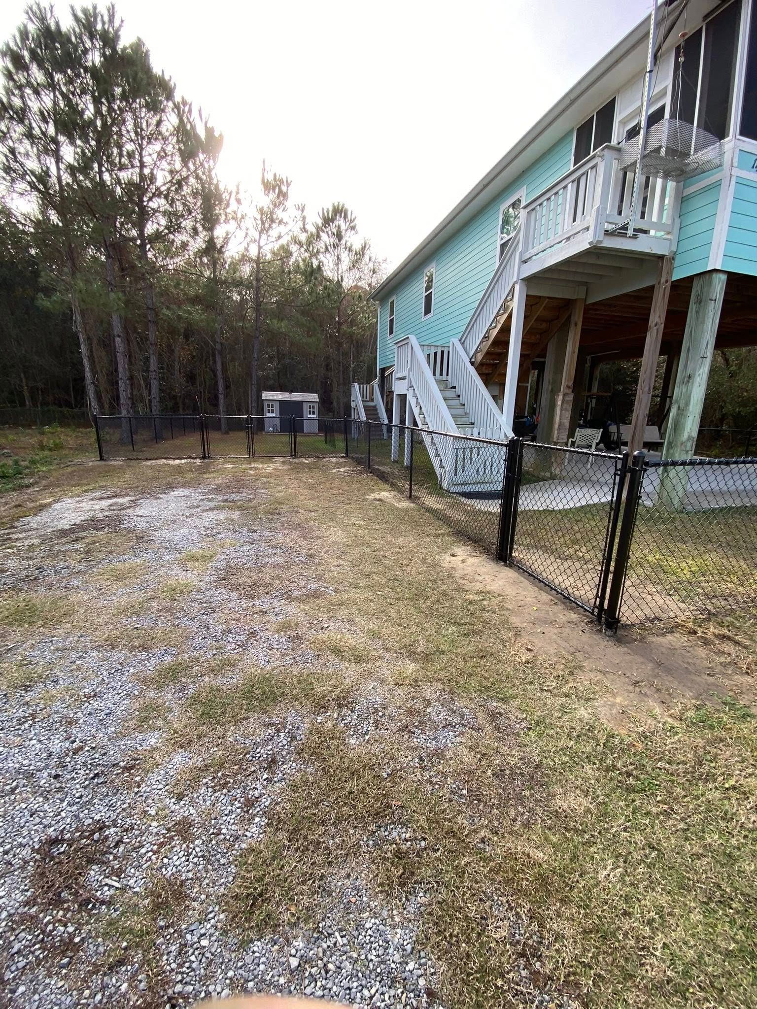 A light blue house on stilts with white stairs, a chain-link fence, and a gravel yard near a wooded area.