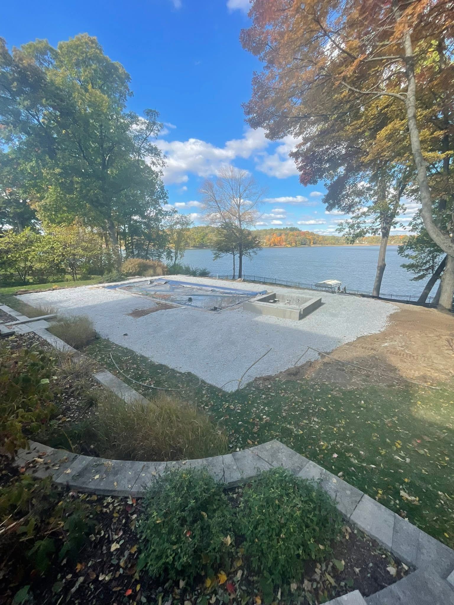 A lakeside backyard with a rectangular gravel patio under construction, bordered by stone steps and autumn trees.