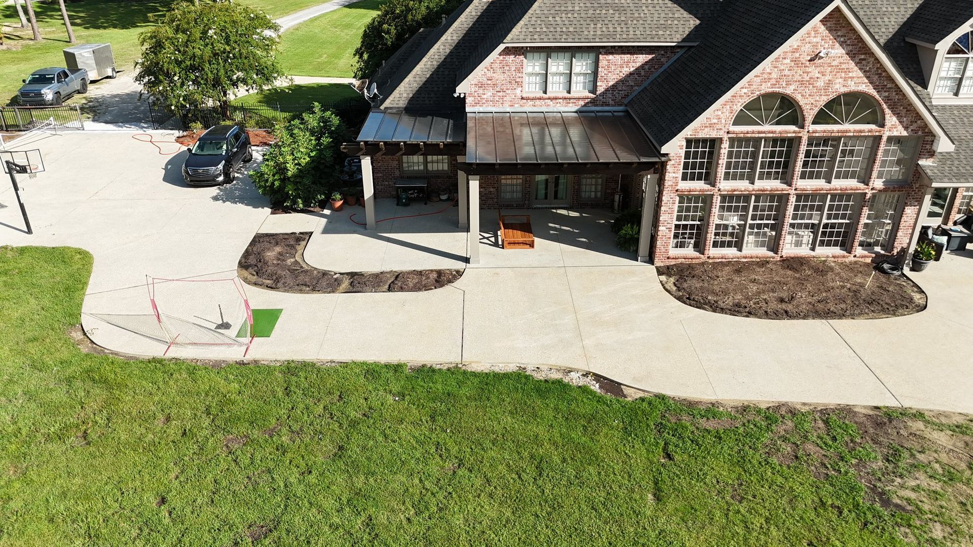 Aerial view of a brick house with a large concrete patio, landscaping beds, and a driveway with parked vehicles.