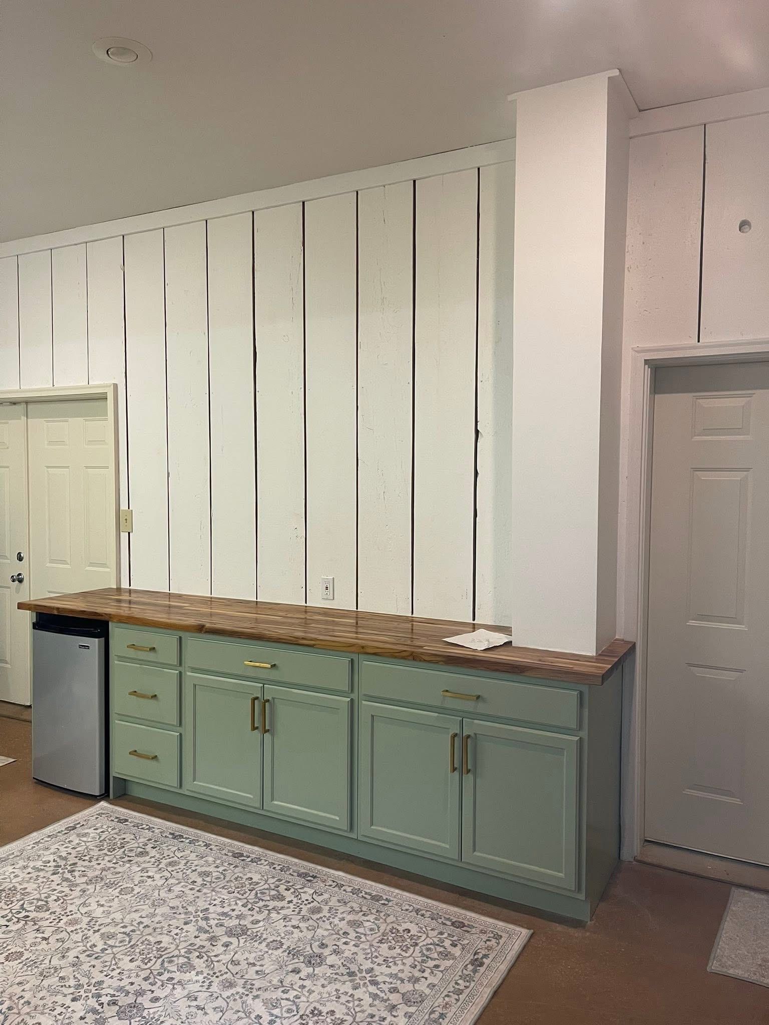 Light green kitchen cabinets topped with a brown counter sit against a white wall with vertical wood paneling.