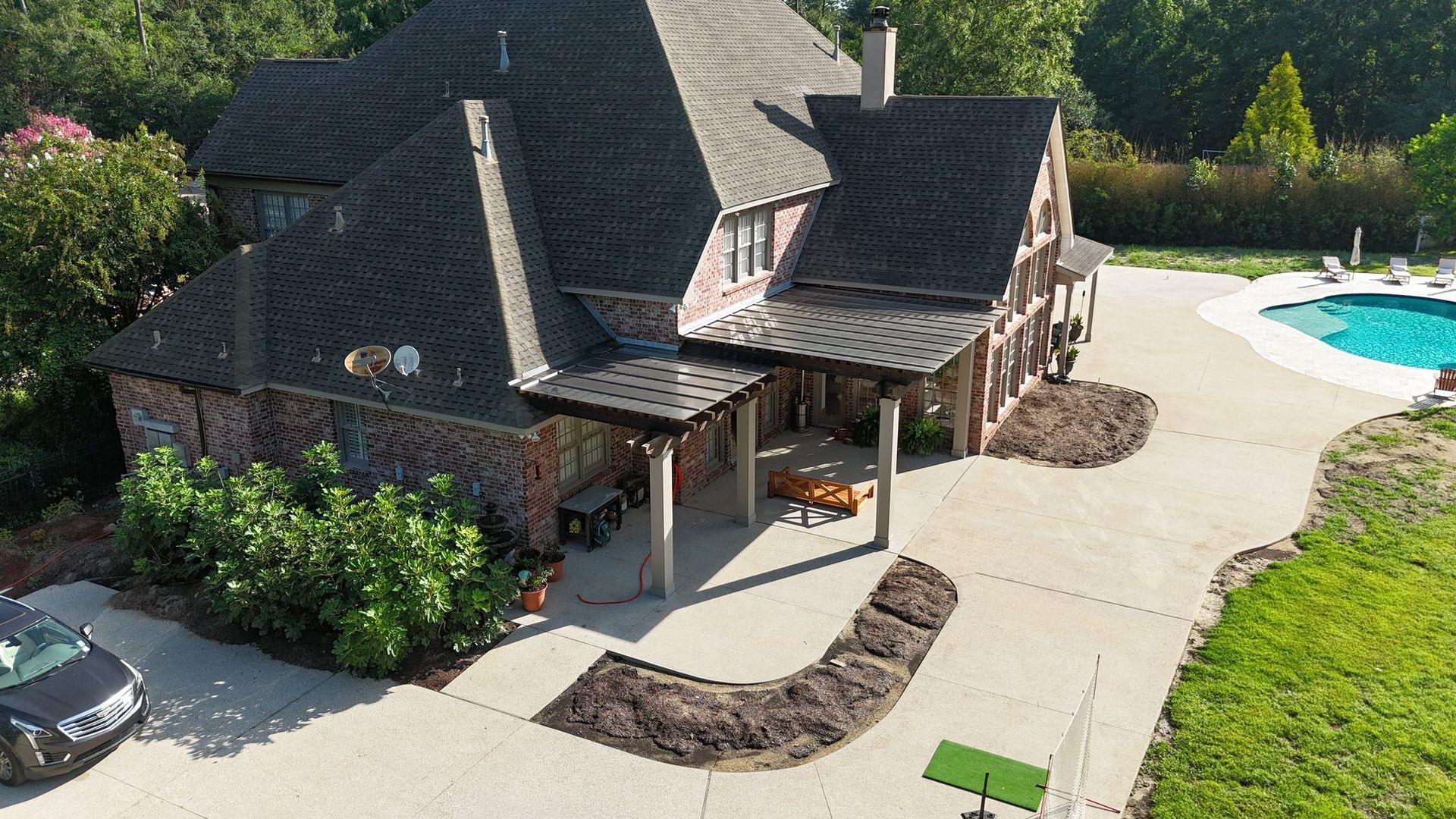 Aerial view of a brick house with a covered patio, adjacent driveway with a car, and an outdoor swimming pool nearby.