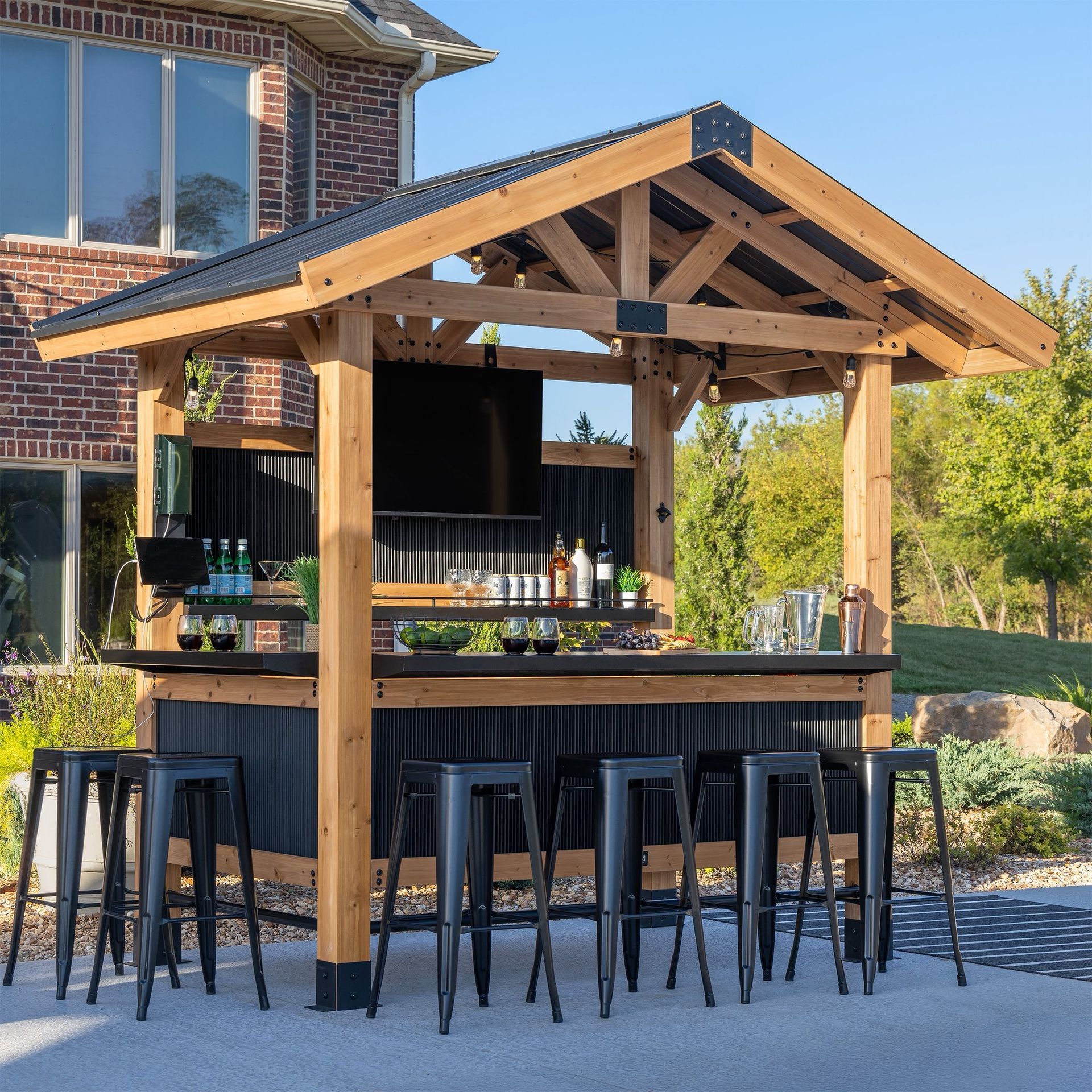 A rustic, open-air wooden outdoor bar with a black corrugated roof, metal paneling, and four black bar stools on a patio.