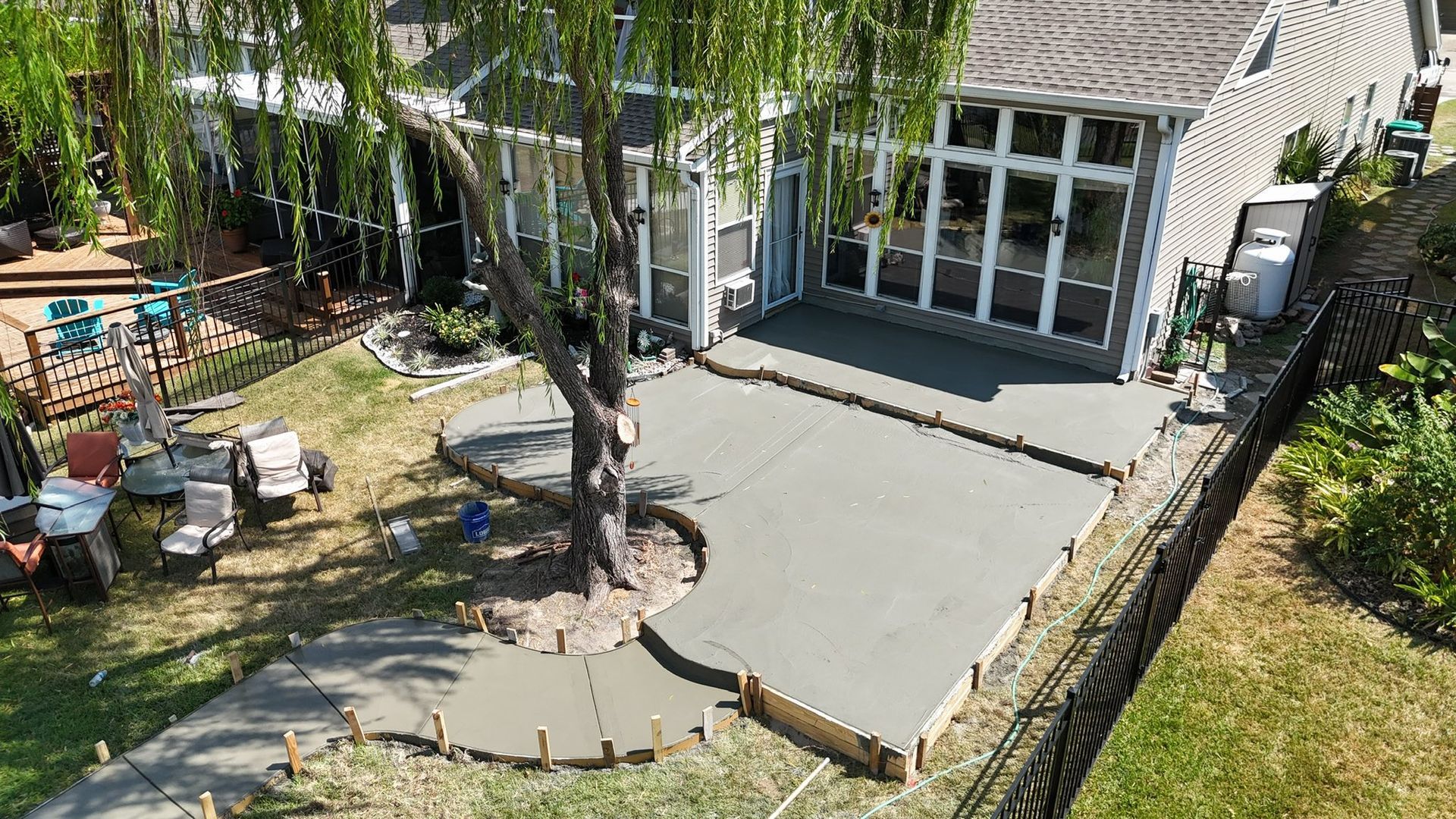 High-angle view of a newly poured concrete patio in a backyard, featuring a curved cutout around an existing tree.