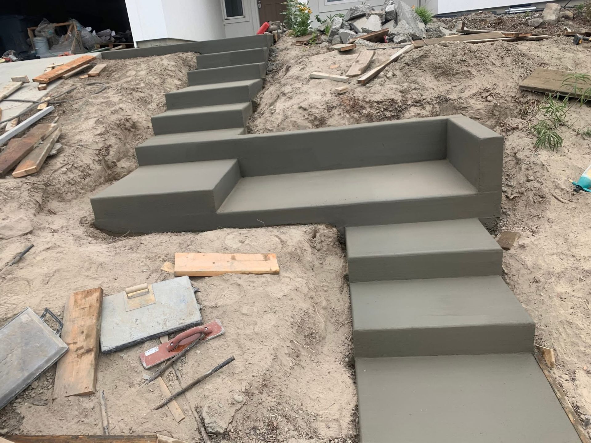 A set of freshly poured concrete stairs and a bench landing under construction on a dirt slope near a building.