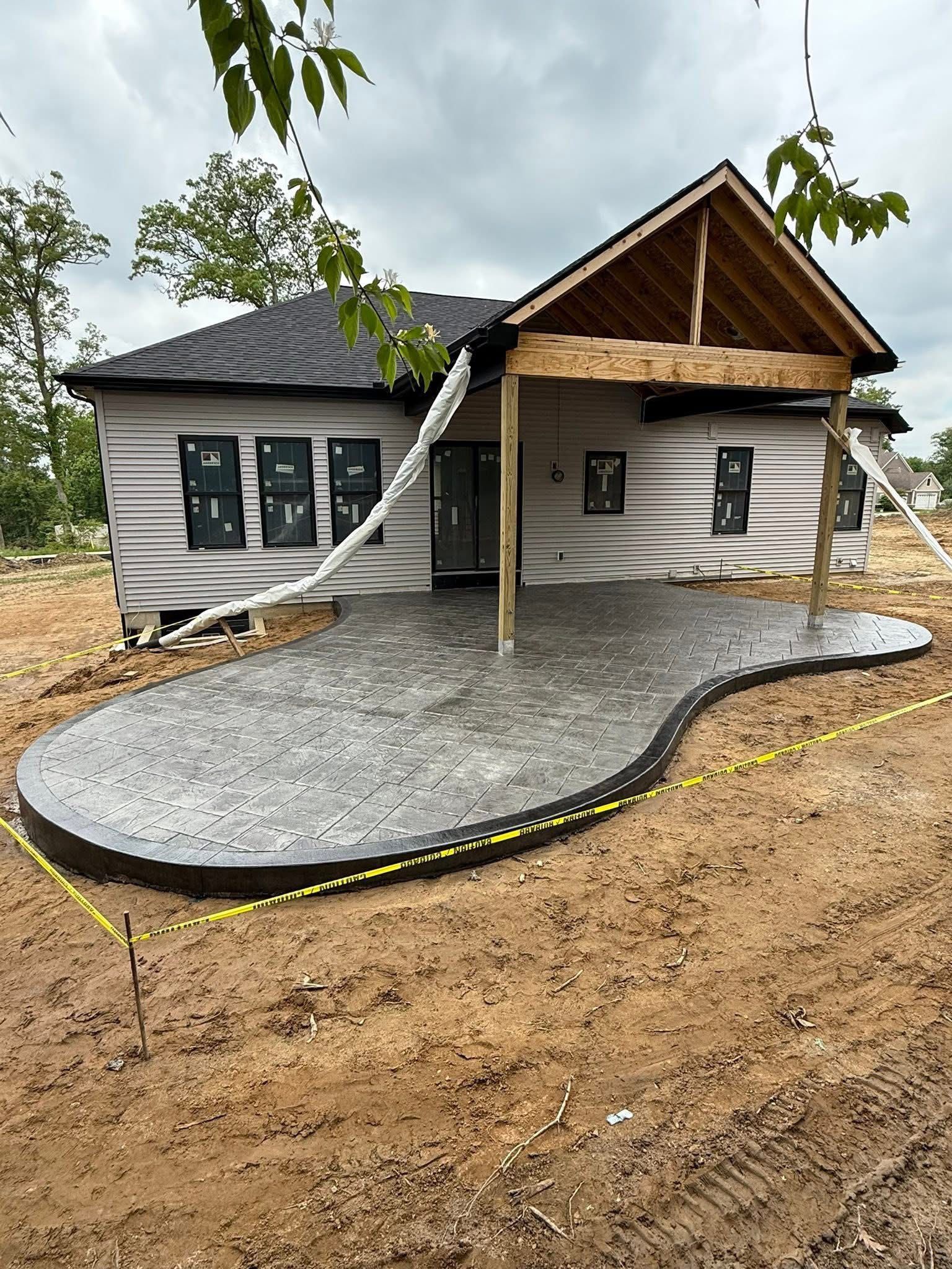 A new house under construction features a large, kidney-shaped stamped concrete patio and an unfinished wooden porch roof.