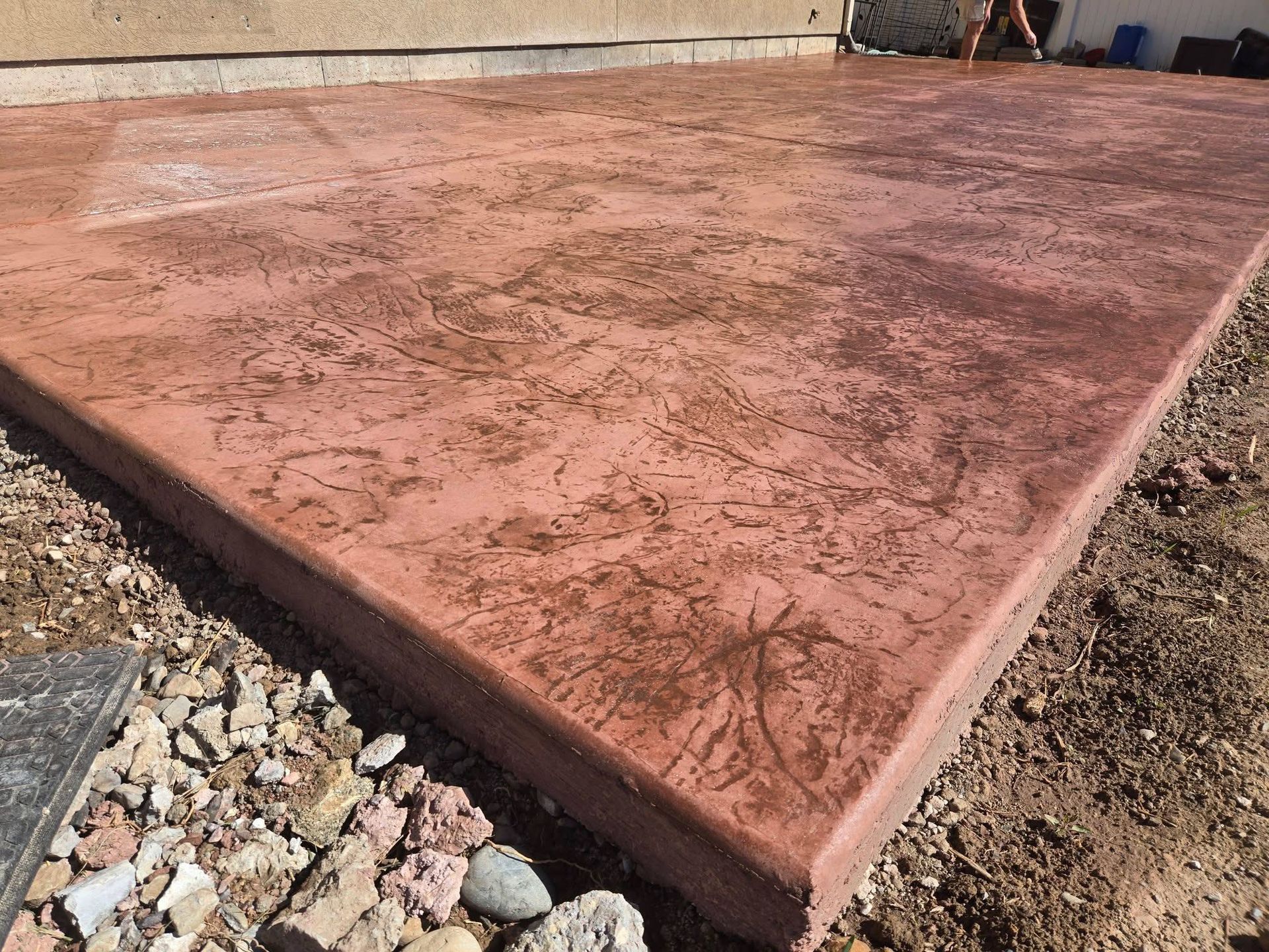 A rectangular, red stamped concrete patio slab sits adjacent to a gravel yard and a stucco wall.