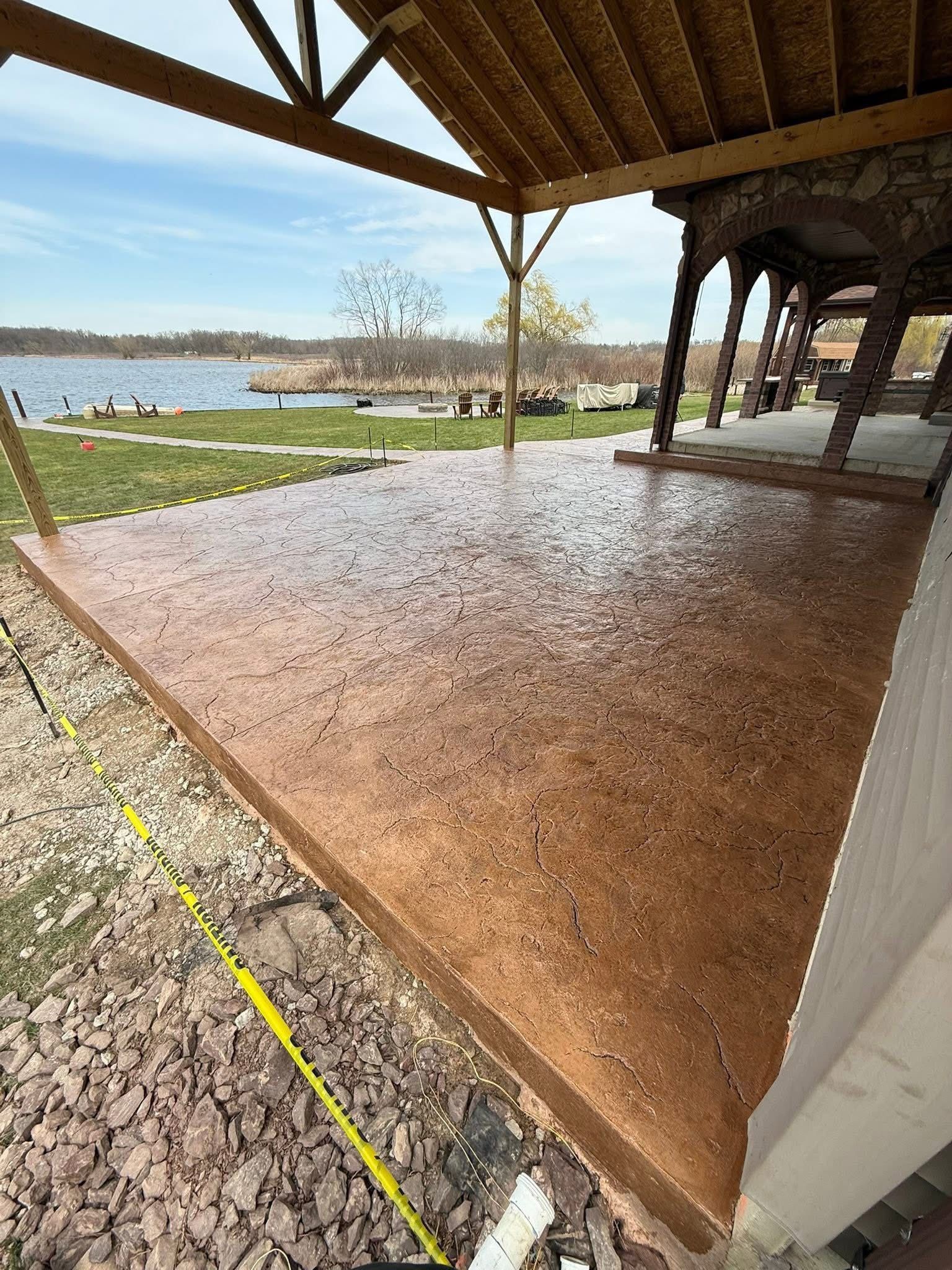 A covered patio with a textured, speckled brown floor overlooking a lake and grassy yard on a sunny day.
