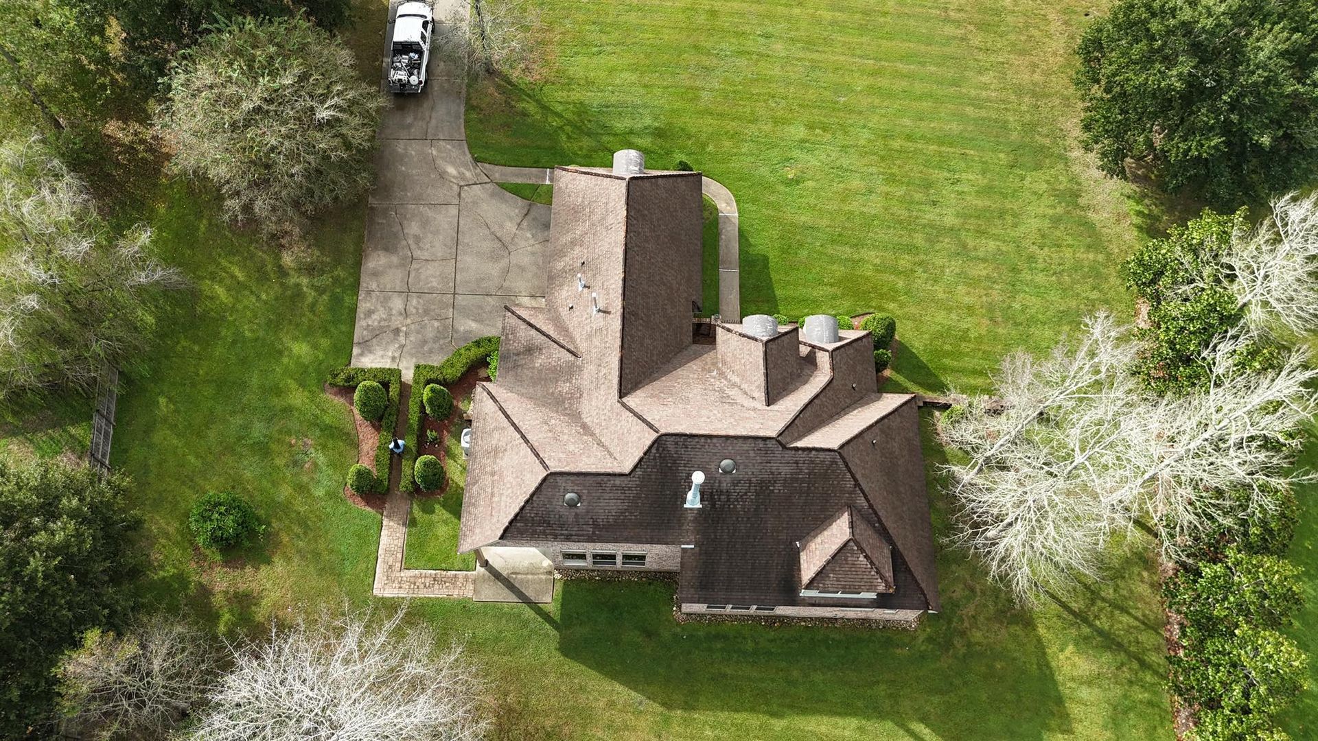 Aerial view of a house with a brown shingled roof, a paved driveway with a parked vehicle, and lush surrounding trees.