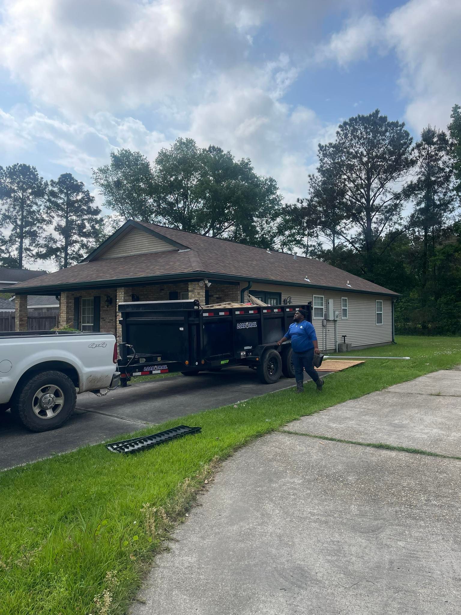 A person in blue stands next to a black dump trailer attached to a white truck parked in front of a house.
